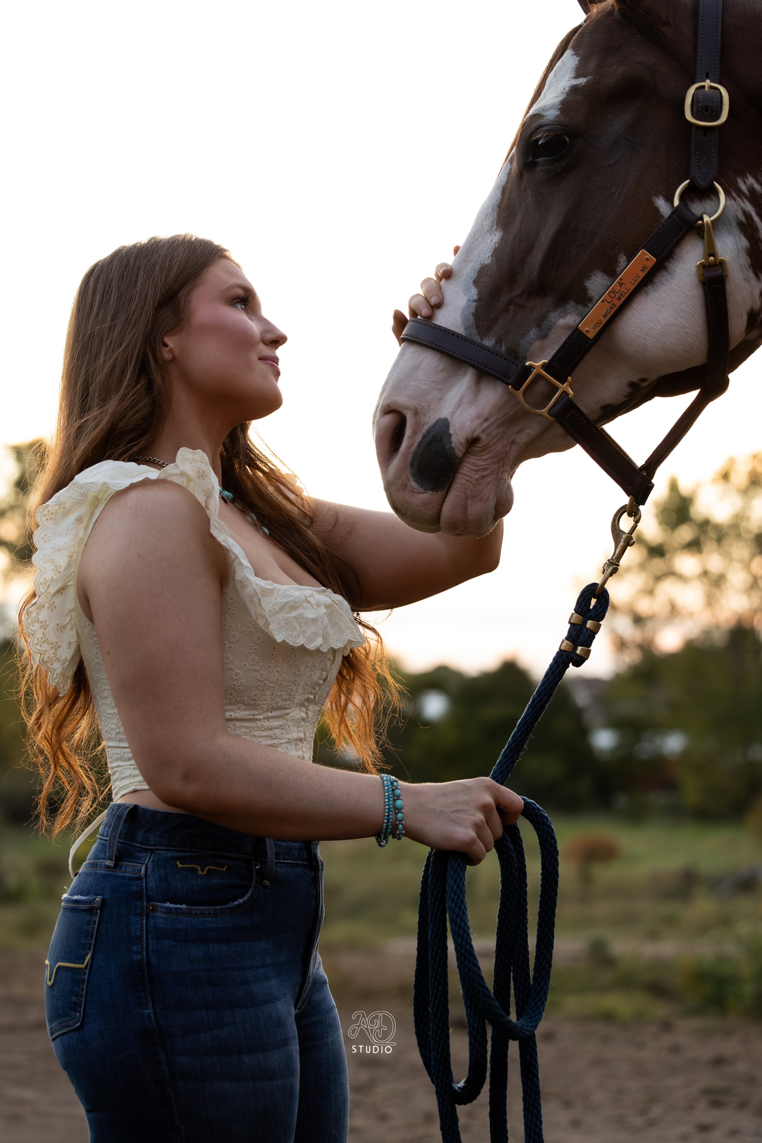 A woman with long brown hair, wearing a light-colored blouse and blue jeans, holding a lead rope while petting the face of a brown and white horse during sunset.