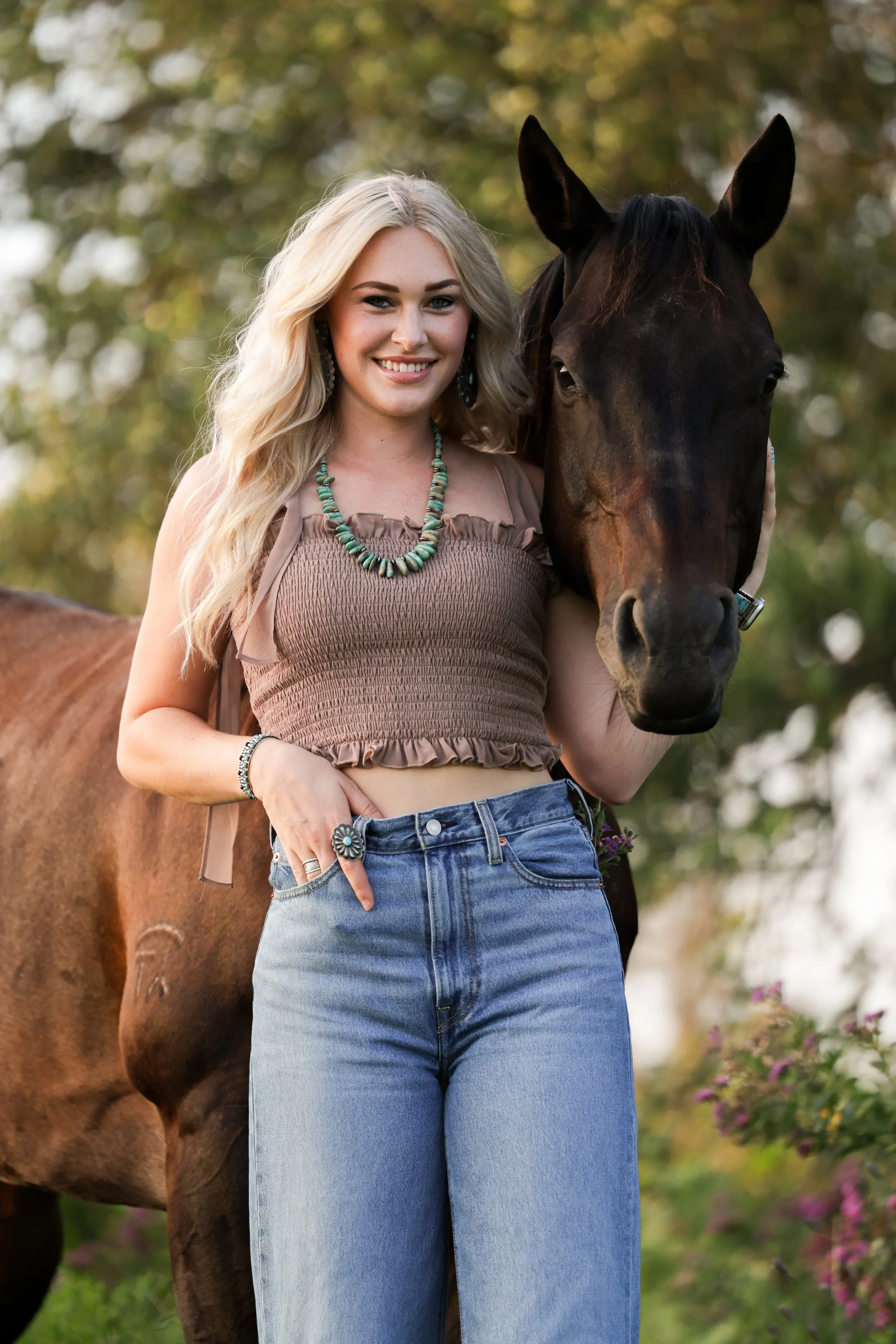 A young woman with long blonde hair and wearing a sleeveless beige top with ruffles, blue jeans, turquoise jewelry, standing outdoors with a brown horse and trees with green and yellow leaves in the background.