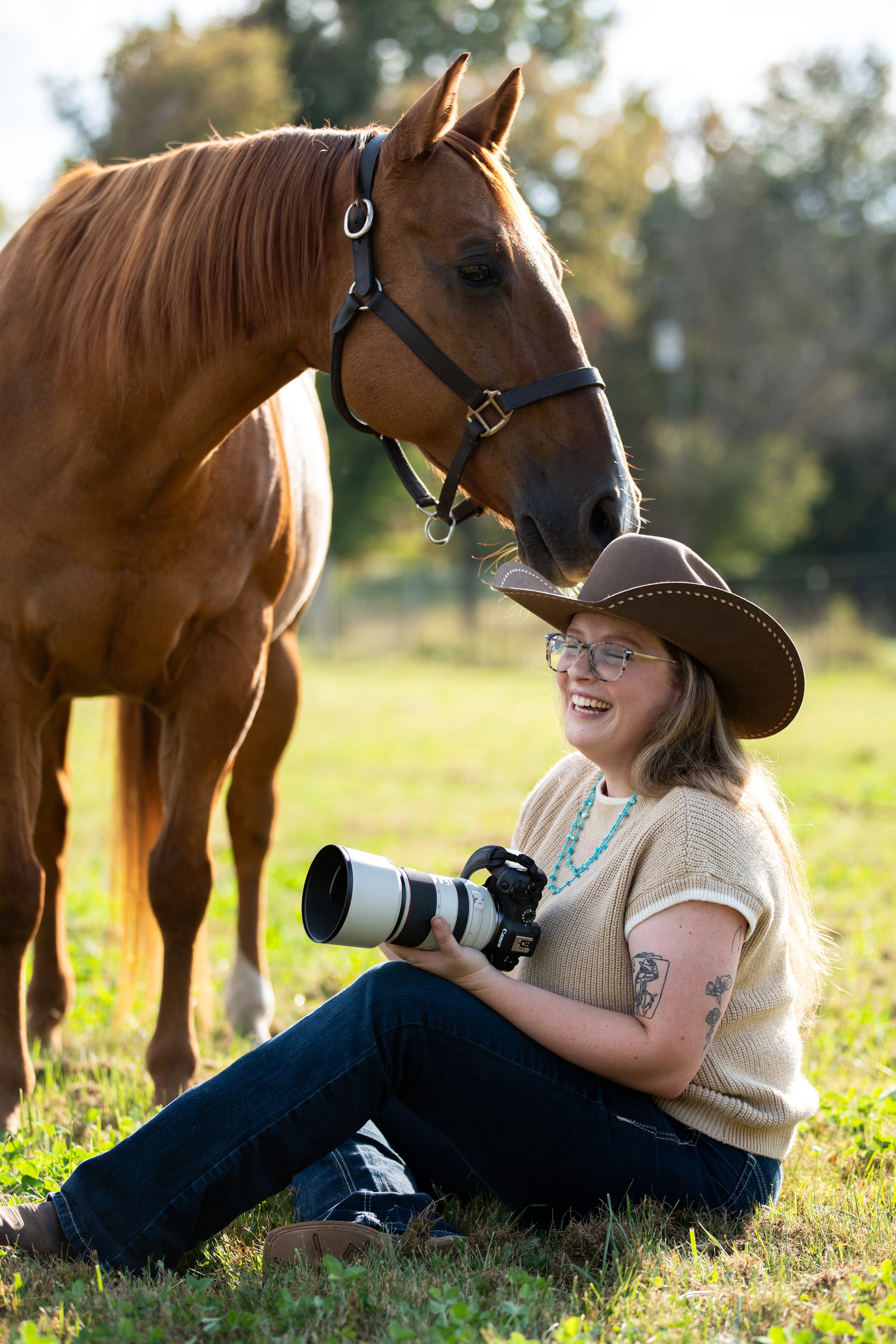 A woman with tattoos on her arms sitting on grass, holding a camera with a large telephoto lens, smiling, wearing glasses, a tan sweater, jeans, and a wide-brimmed hat, with a horse standing close to her, outdoors with a blurred background of trees and sunlight.