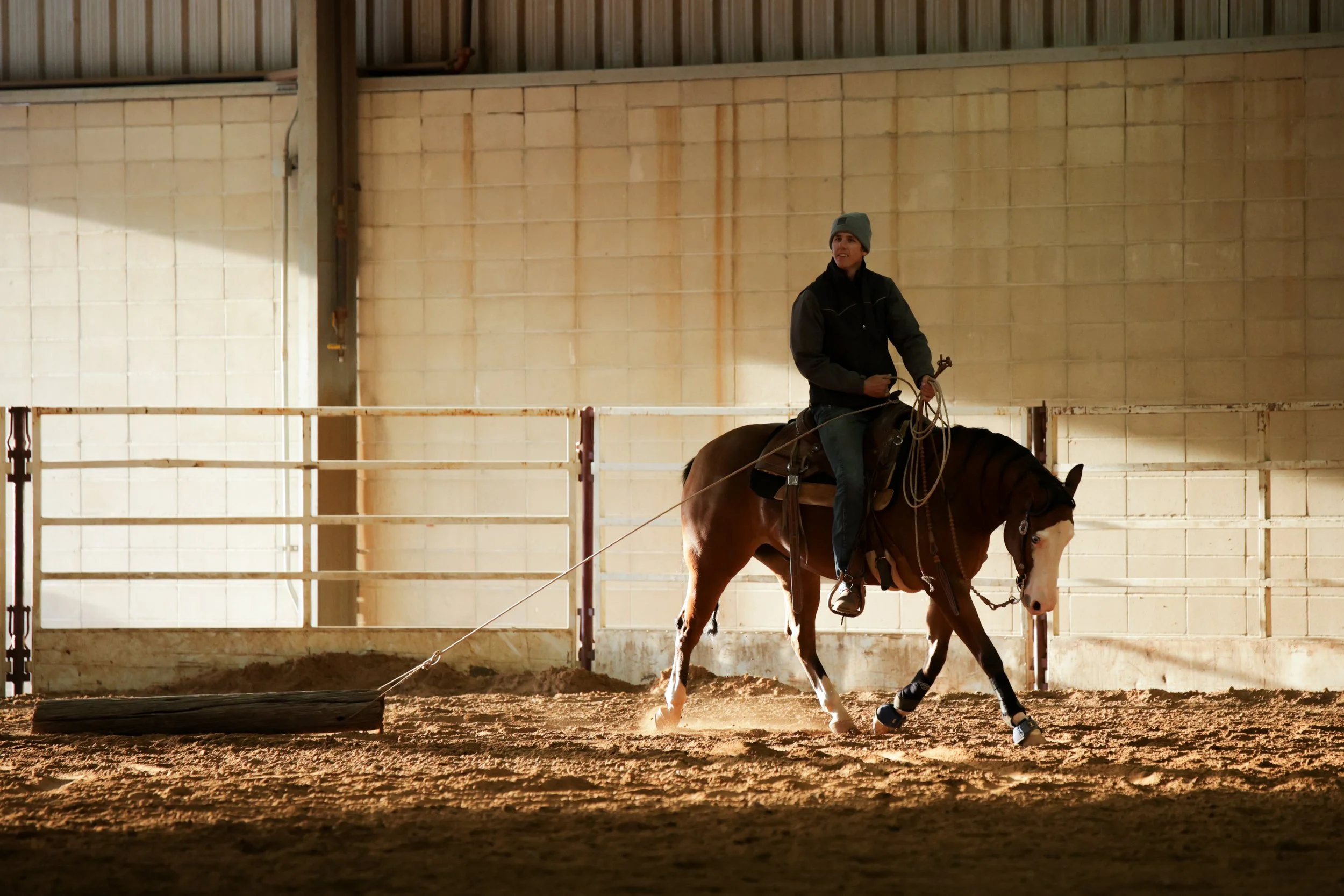 Person riding a horse inside a riding arena while dragging a log.