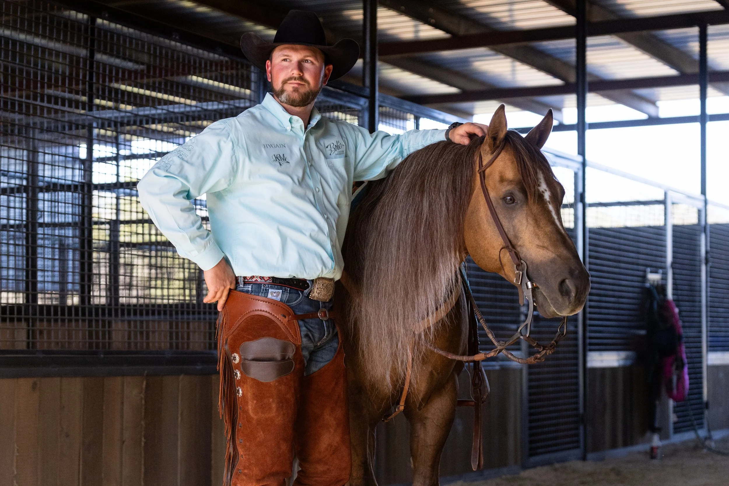 A man wearing a cowboy hat and a light blue shirt standing beside a brown horse inside a barn.