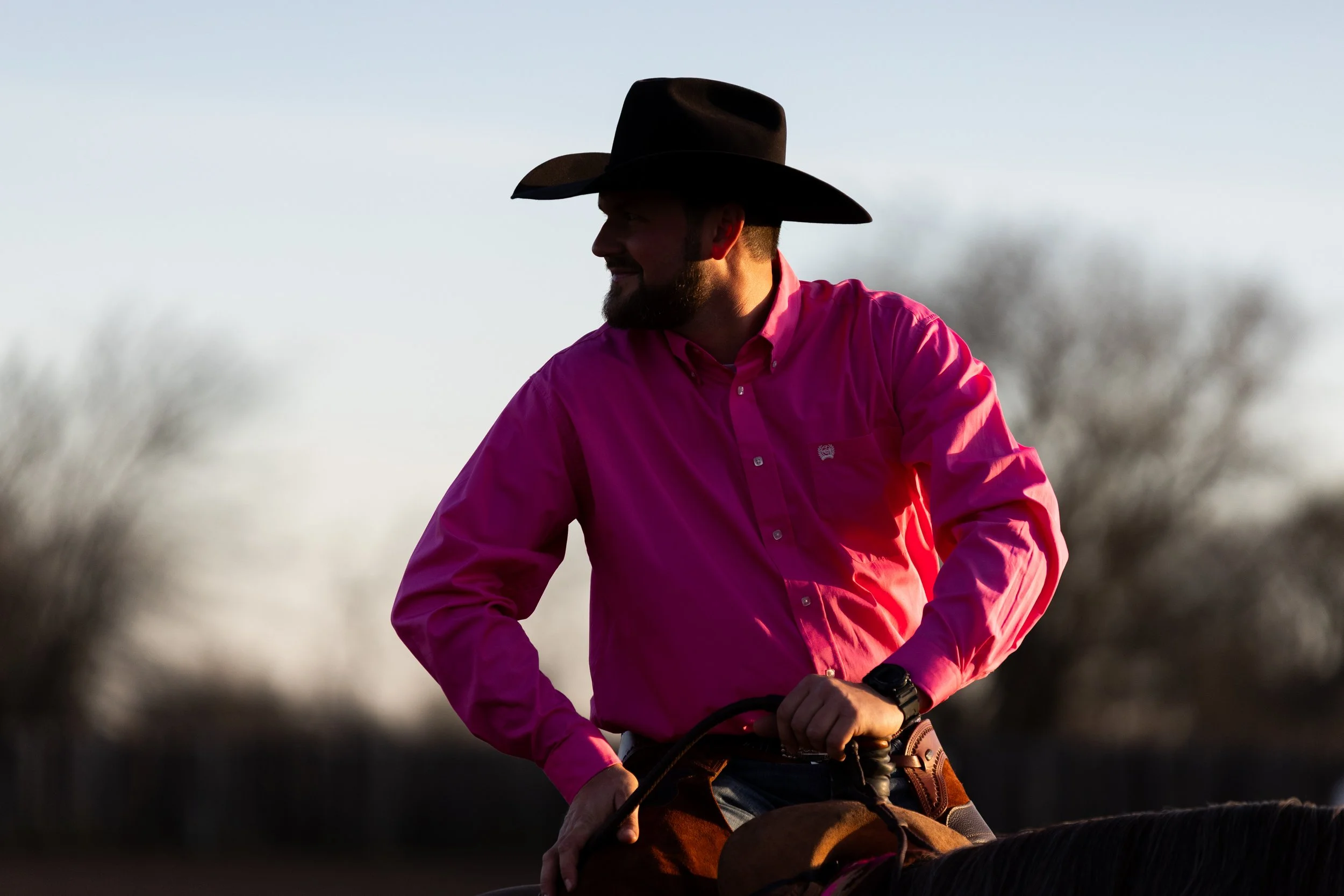 A man wearing a black cowboy hat and a bright pink shirt is riding a horse outdoors during sunset, with blurred trees in the background.