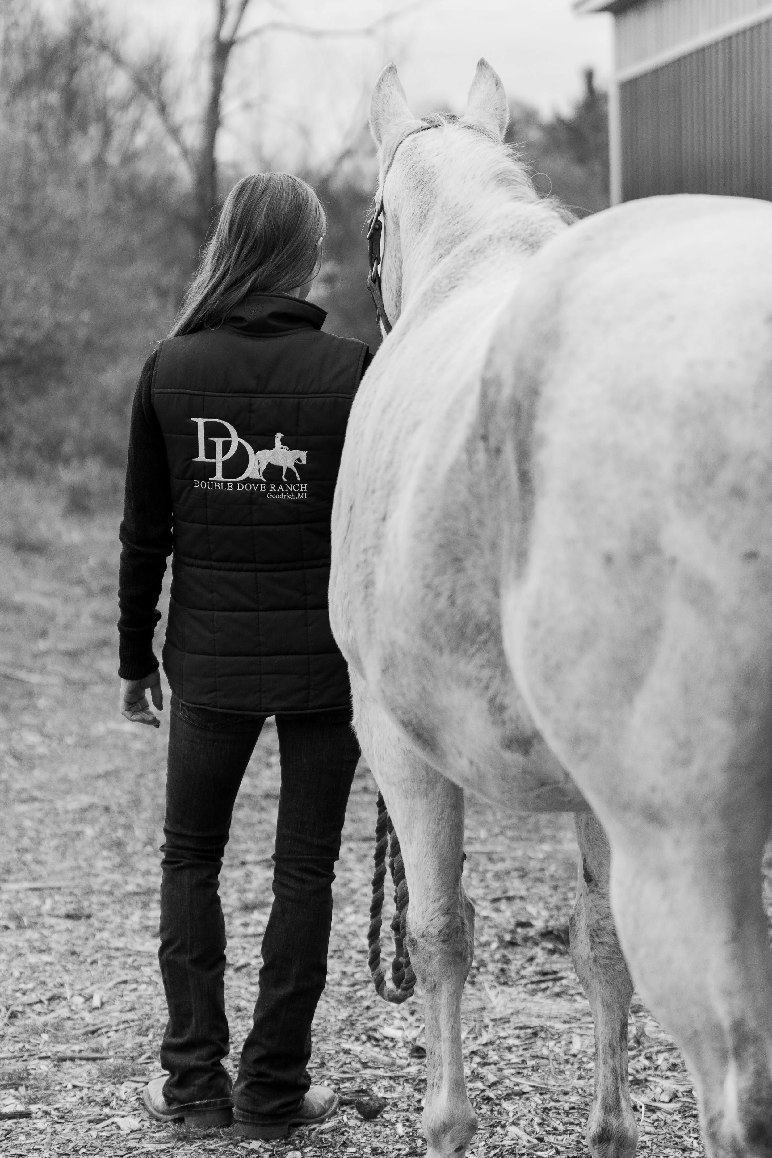 A black and white photo of a person standing next to a white horse outdoors, seen from the back. The person is wearing a vest with the logo and name of Double Dove Ranch in Goodrich, MI.