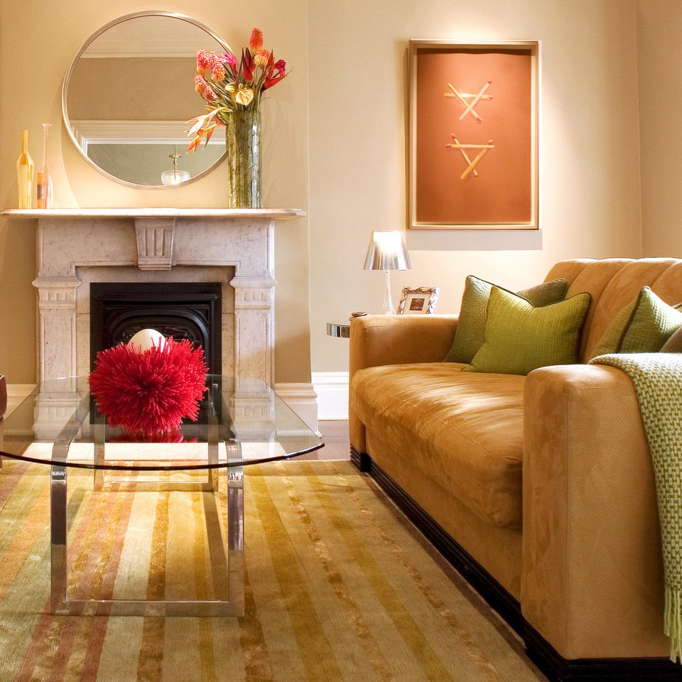 Living room with a beige sofa, green pillows, a glass coffee table, a fireplace with a marble mantel, a large round mirror, colorful vases, a tall flower arrangement, a framed abstract artwork, a table lamp, and a colorful area rug.