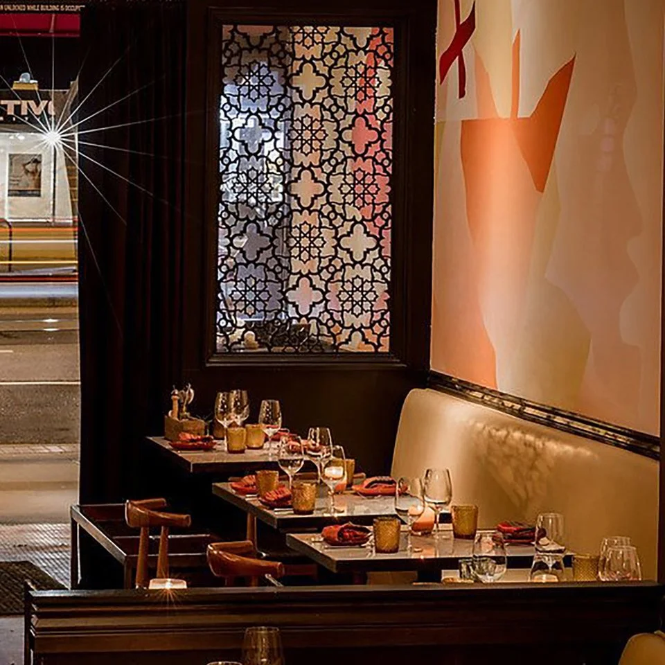Empty restaurant table set with wine glasses, candles, and folded napkins, next to a painted wall with orange and peach hues and a decorative window with floral lattice pattern, outside view shows a city street with passing cars.