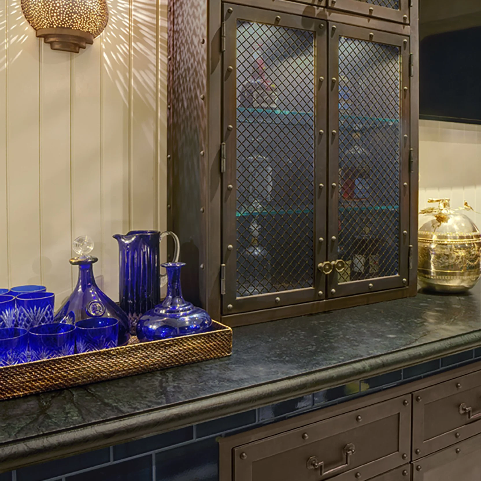 A display of blue glassware including pitchers, vases, and glasses on a basket tray on a kitchen countertop, with a metal cabinet behind and a decorative gold urn to the right.