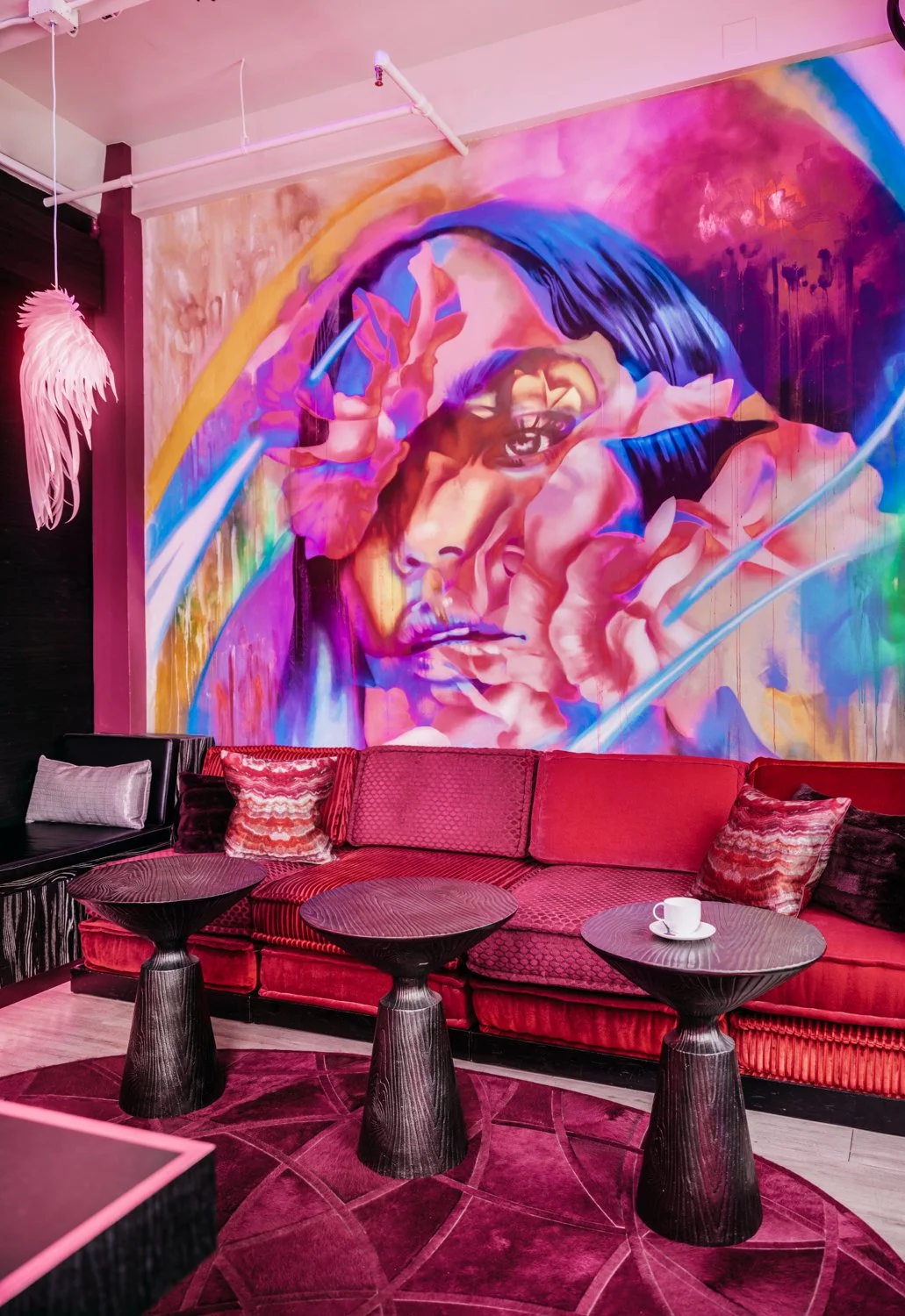 A colorful mural of a woman with short hair making a gesture with her fingers near her eye, on a vibrant pink and purple background. The room has a red sofa with patterned cushions, three dark wood tables, a pink feathered chandelier, and a purple and pink geometric carpet.