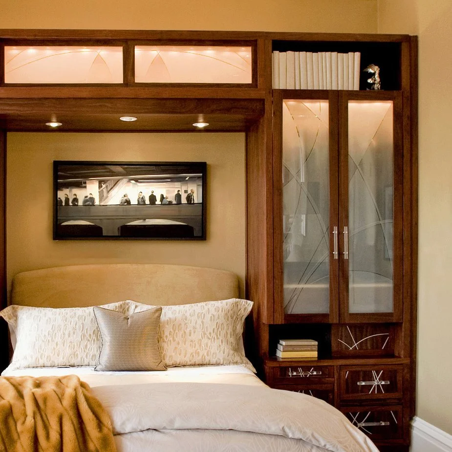 A bedroom with a headboard, decorative pillows, and a blanket. Over the bed is a framed black-and-white photo of people on an escalator in a subway station. To the right, there is a wooden cabinet with glass doors, books, and decorative items.