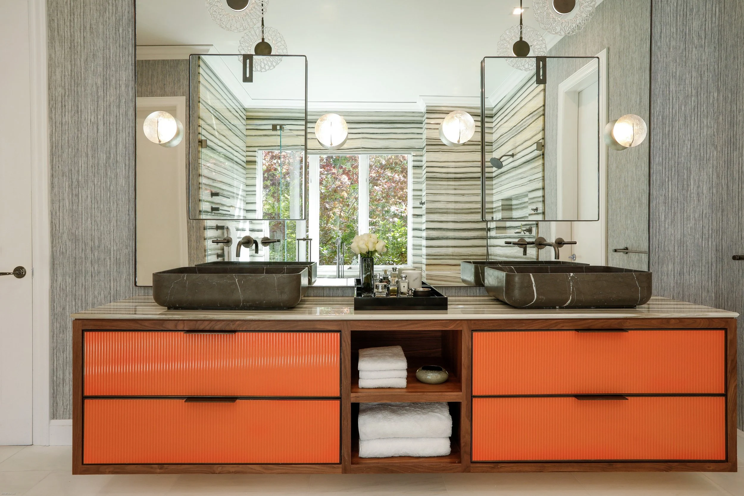 Modern bathroom vanity with two black vessel sinks, a black tray with toiletries, a flower vase with white flowers, and towels stored in open compartments, set against a wall with horizontal striped marble tiles and a large mirror, with a window showing trees outside.