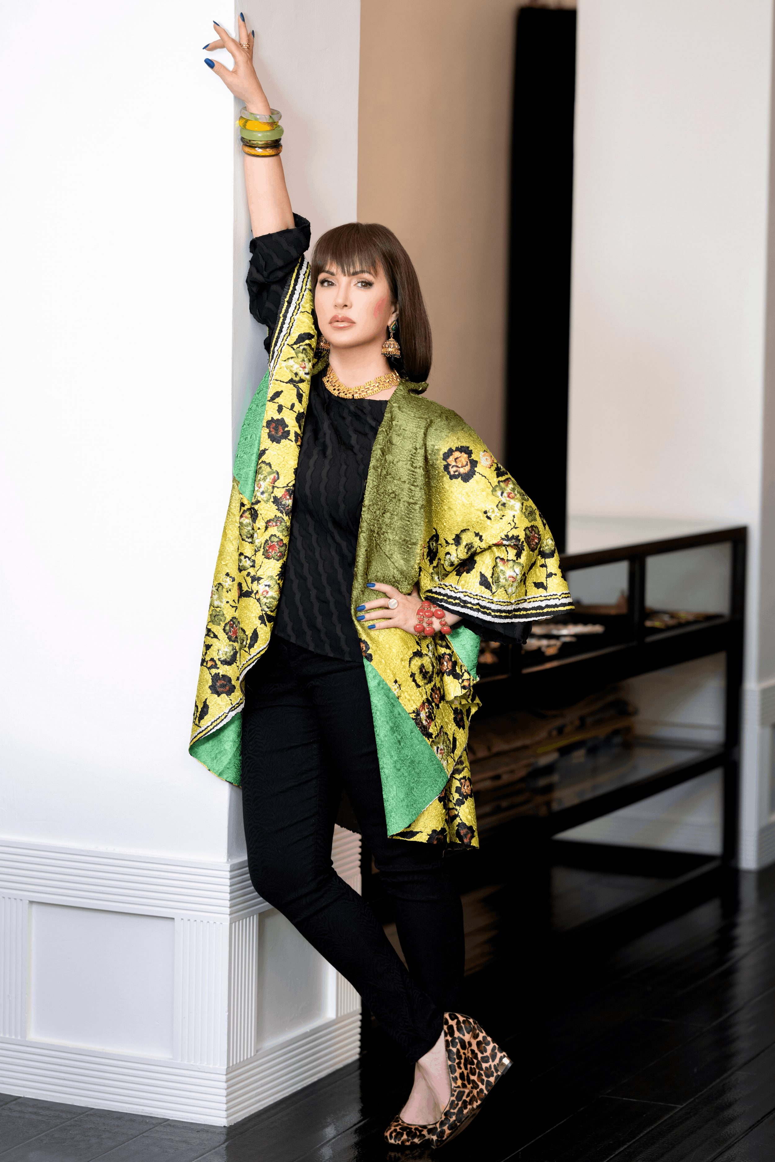 A woman with shoulder-length brown hair and bangs standing against a white wall. She's wearing a colorful, patterned coat with yellow, green, and black details over a black top and black pants. She accessorizes with large earrings, a gold necklace, multiple bracelets, and leopard-print high heels. She's posing with her right arm raised, her hand touching the wall, and her left hand on her hip.