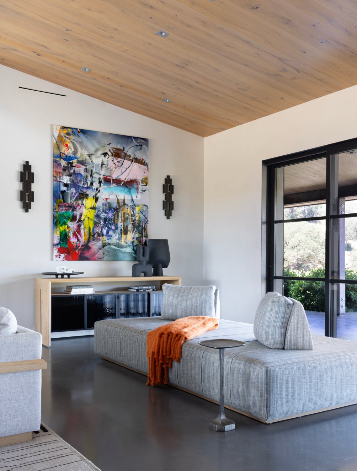 Modern living room with light gray sofa, orange throw, abstract colorful painting on white wall, black sideboard, and large windows showing outdoor greenery.
