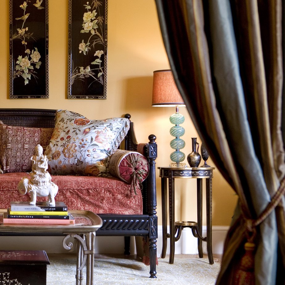 Close-up of a cozy living room corner with black wooden furniture, a floral pillow, a small figure sculpture on a tray, and a side table with a patterned lamp and decorative vases, partly obscured by a curtain.
