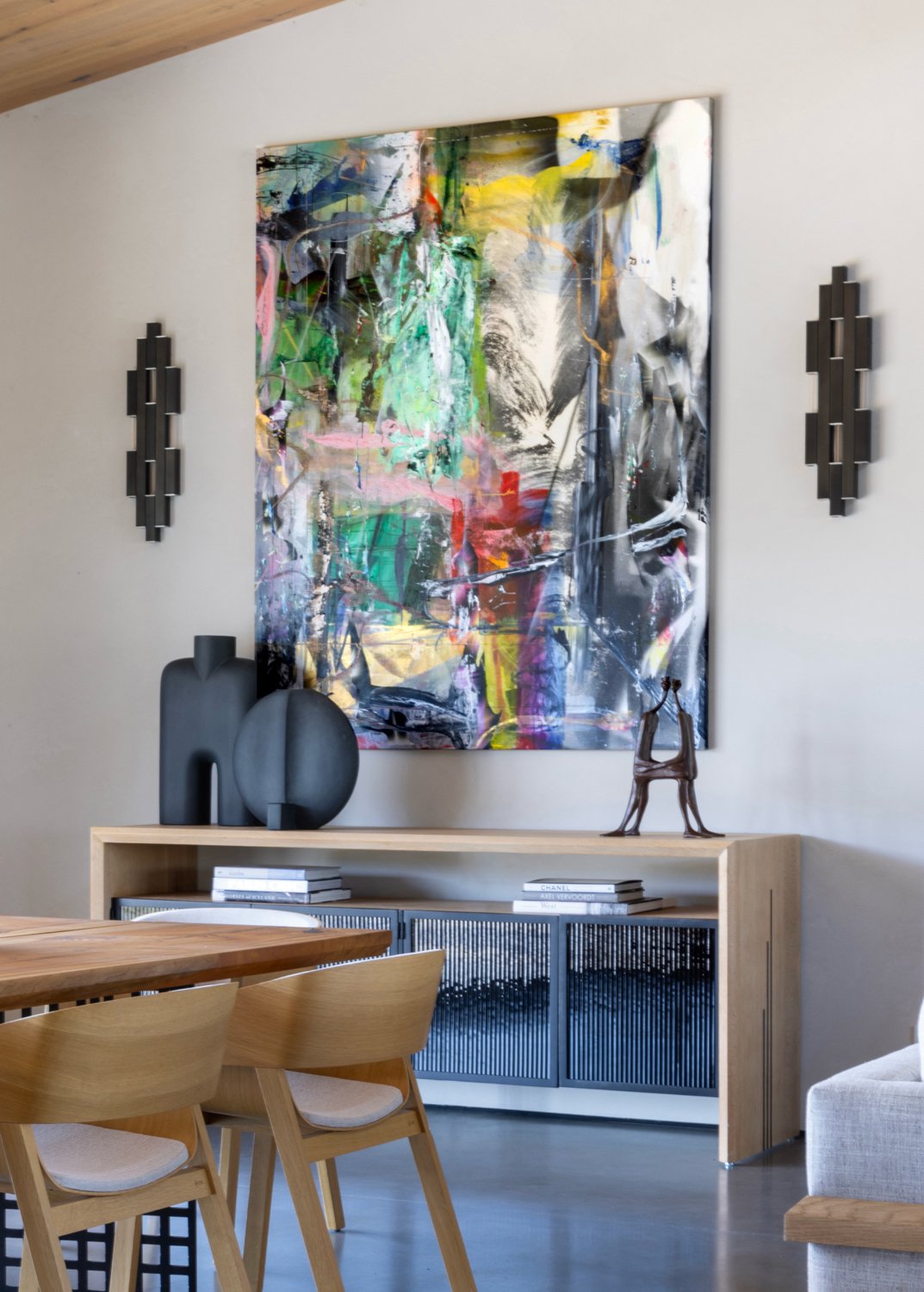 Interior of a modern living space featuring a large colorful abstract painting on the wall, matching wall sconces, a wooden sideboard decorated with black vases and a sculpture, and surrounding furniture including a wooden table and chairs.