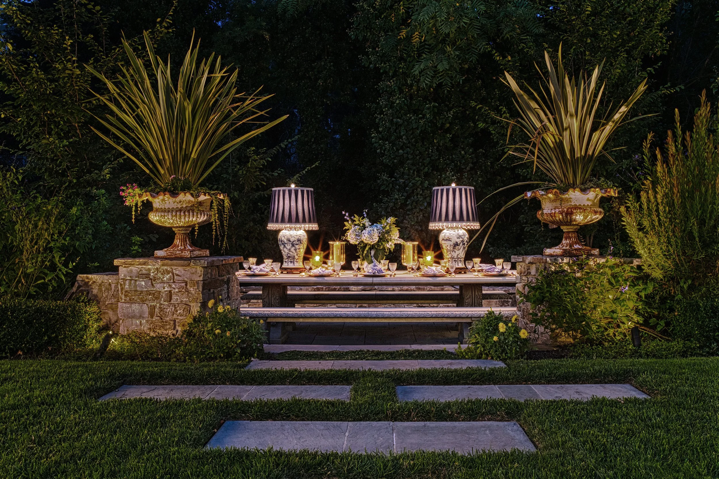 Outdoor evening scene of a dining area with a long wooden table set for a meal, surrounded by large decorative urn planters with tall foliage, illuminated by string lights and table lamps, with a lush green background and a stone pathway leading to the setup.