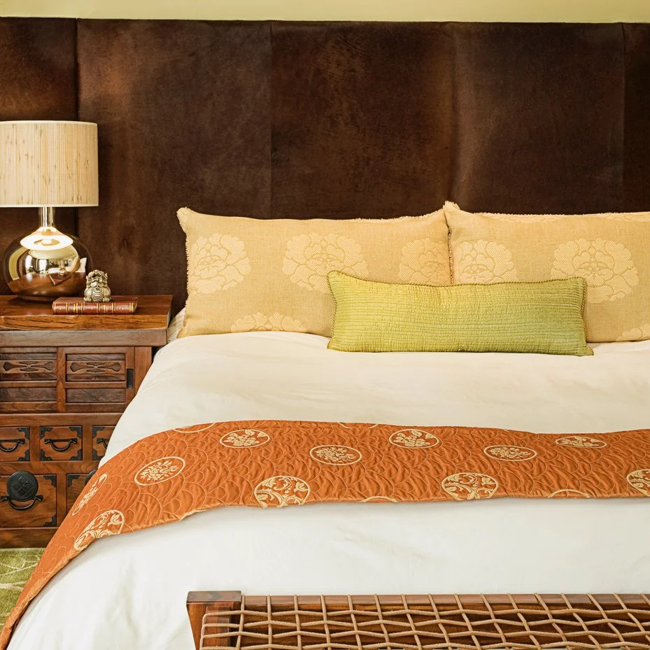 A neatly made bed with beige and green pillows, an orange quilted bed runner, a wooden headboard, and a wooden nightstand with a modern lamp and small decorative items.