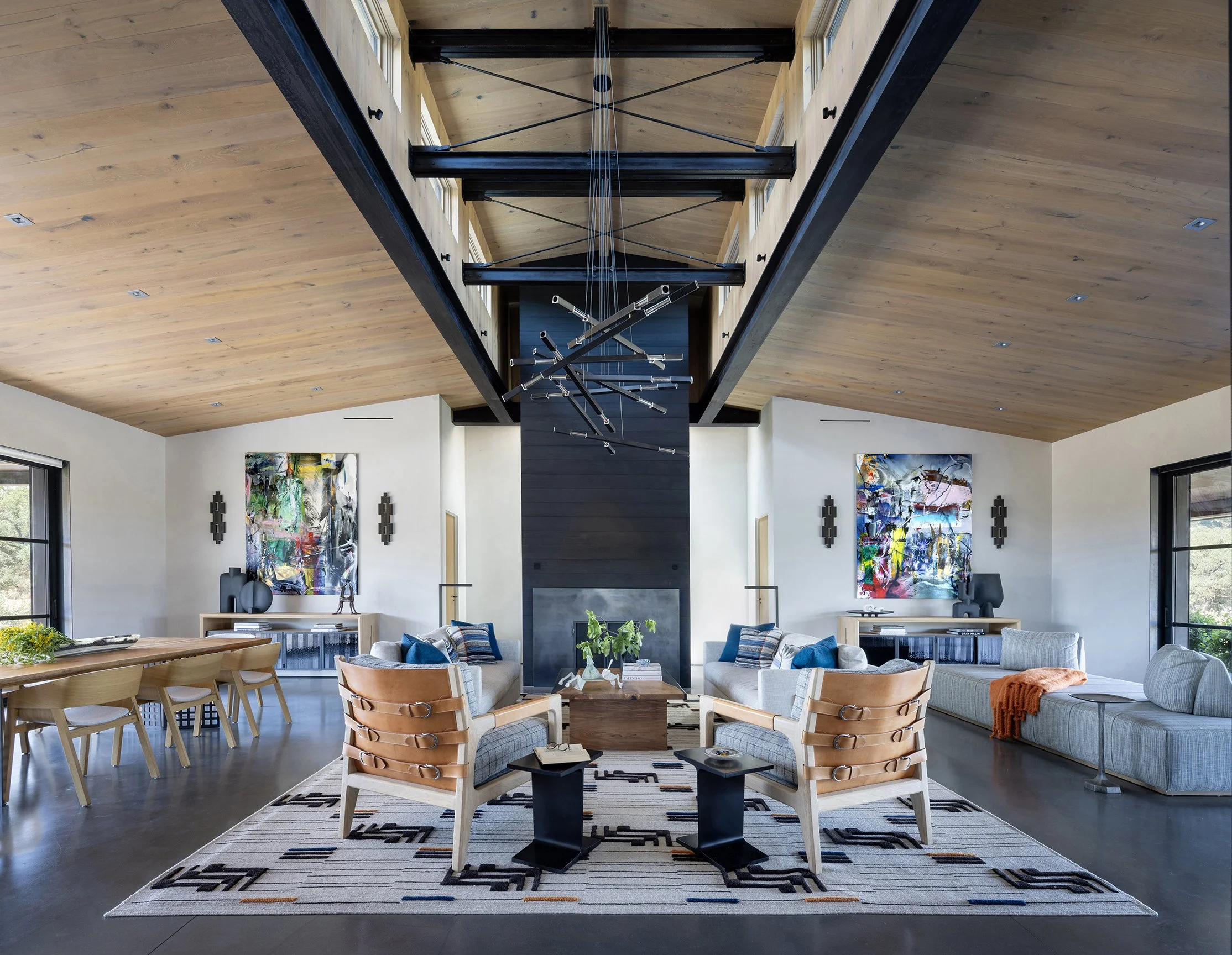Open-concept living room with high wooden ceiling, contemporary chandelier, and large abstract paintings on walls. Furniture includes sofas, armchairs, a wooden dining table with chairs, and decorative items like vases and books. Sunlight streams through large windows.