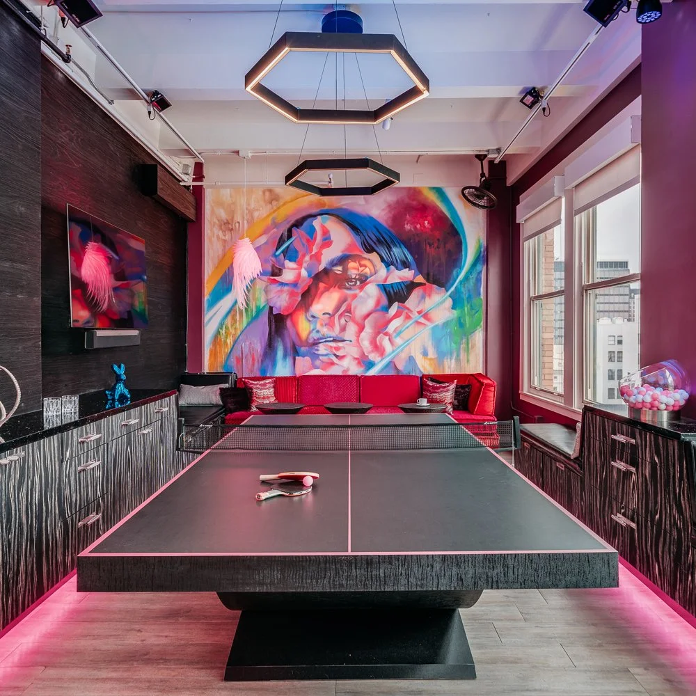 A modern recreation room with a ping pong table in the center, pink lighting underneath, a colorful mural of a woman on the wall behind a red couch, a flat-screen TV on the left wall, large windows on the right side, and decorative pink feathers hanging near the mural.