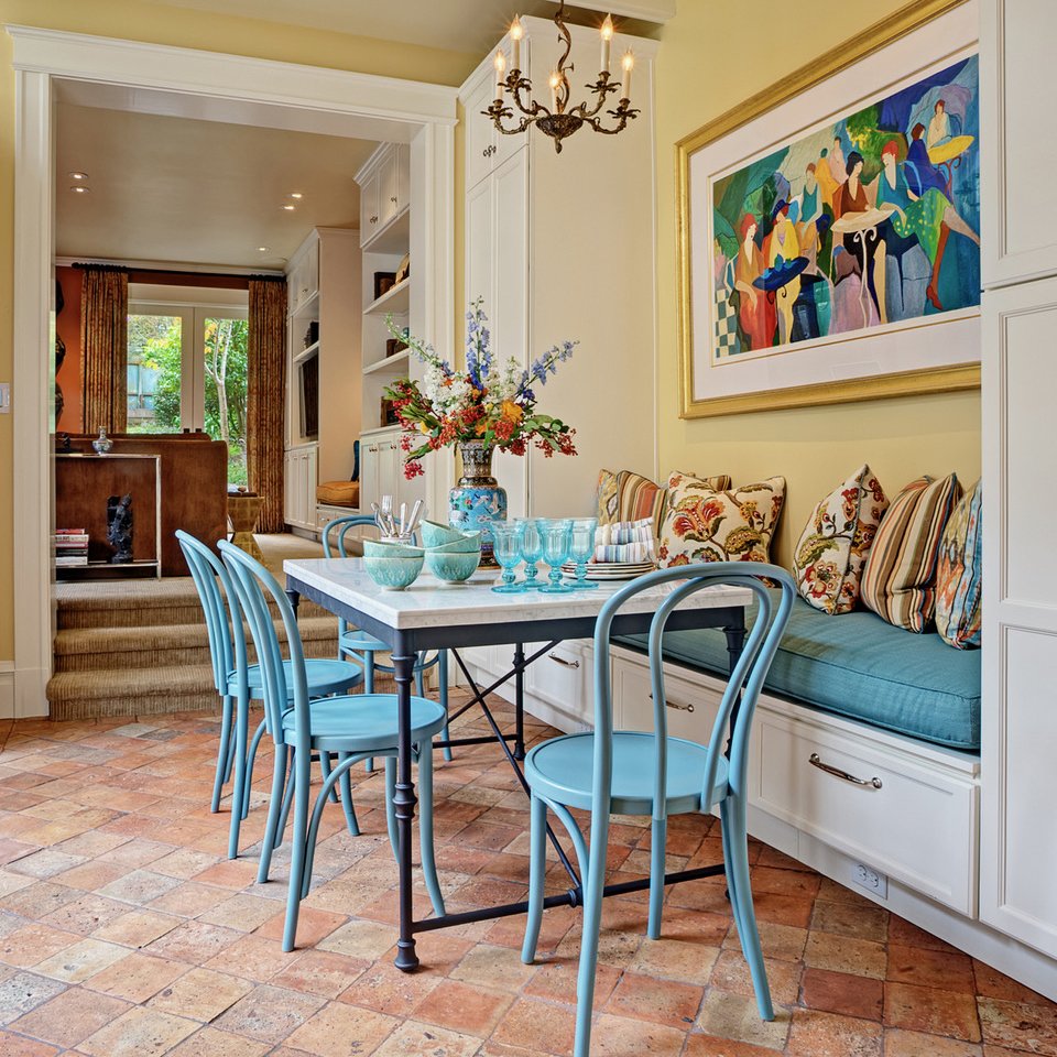 A cozy dining area with a white marble table surrounded by five blue chairs. The table has bowls, glasses, and utensils, and a large colorful flower arrangement in a blue vase. There is a cushioned built-in bench with patterned and striped pillows, and a colorful abstract painting on the wall overhead. A chandelier hangs above the table, and the room opens to a living space with a window and a view of greenery outside.