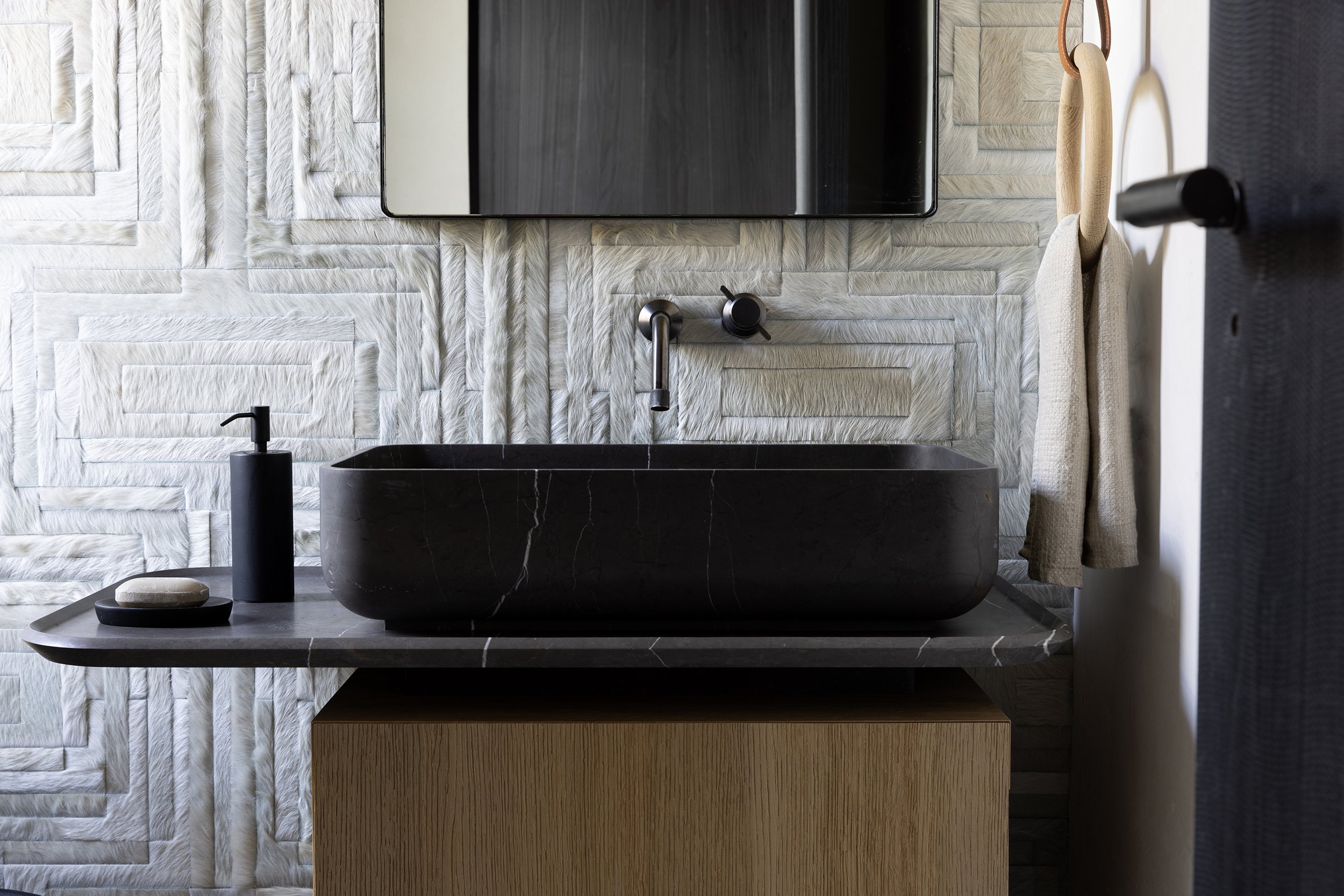 Modern bathroom with textured white wall tiles, a black vessel sink on a gray marble countertop, a wall-mounted faucet, and a mirror mounted above the sink. There is a beige towel hanging on a hook and a black soap dispenser on the countertop.