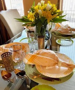 Dining table set with yellow and peach napkins, glassware, and a floral centerpiece of yellow and white flowers.