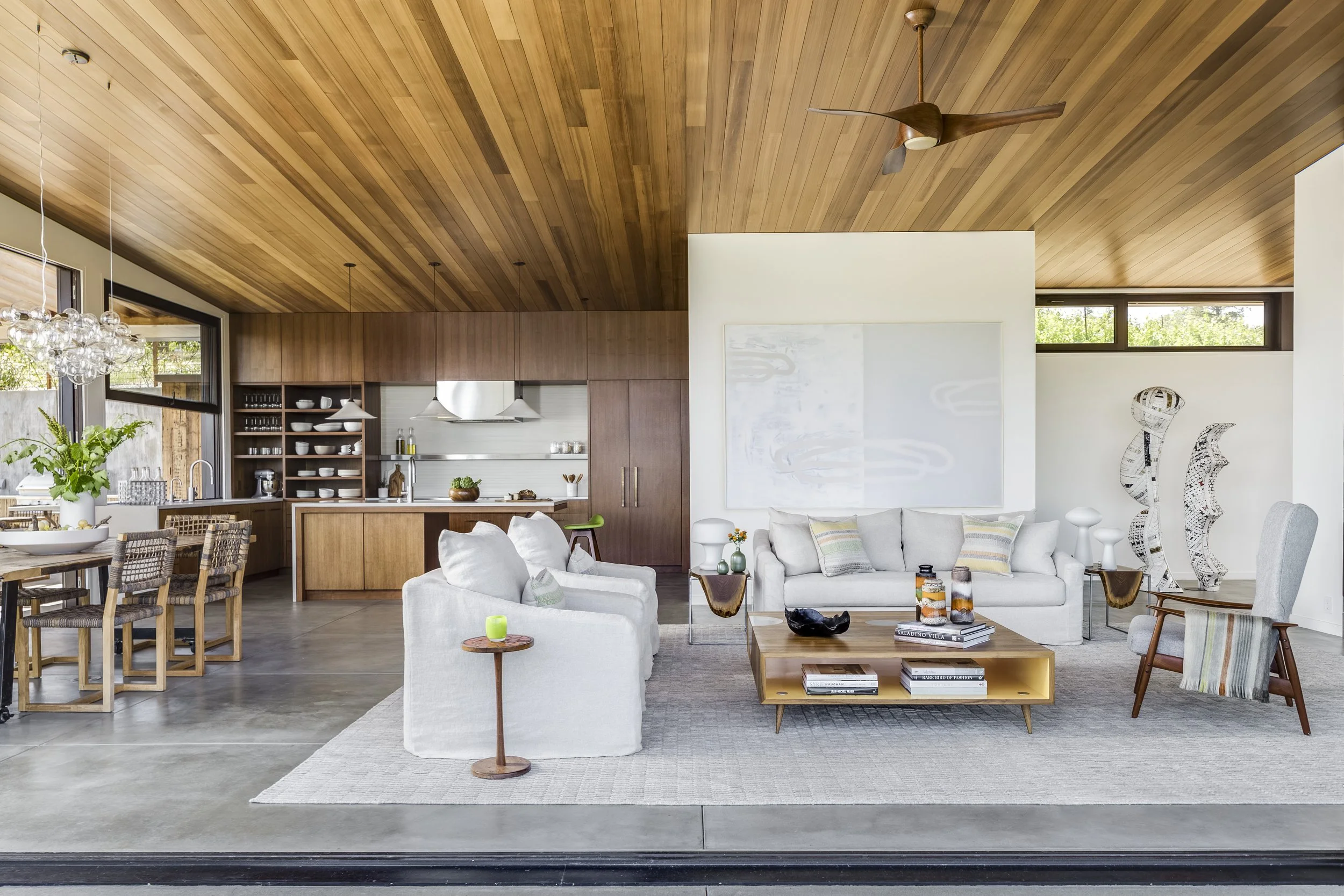 Open-concept living space with a wooden ceiling, white walls, sitting area with white sofas, a wooden coffee table, and decorative objects, adjacent to a kitchen with wooden cabinets and open shelves, and large windows bringing in natural light.