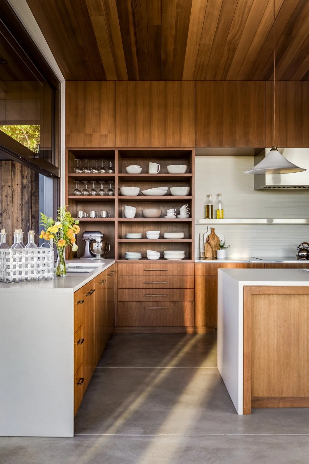 Modern kitchen with wooden cabinets and open shelving, white countertops, a vase of yellow flowers, and white dishes on display.