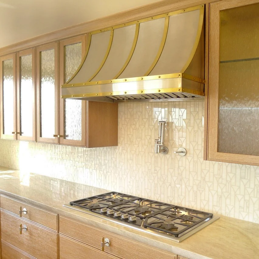 Kitchen with wooden cabinets, a beige backsplash, a gas stove, and a custom gold and beige range hood.