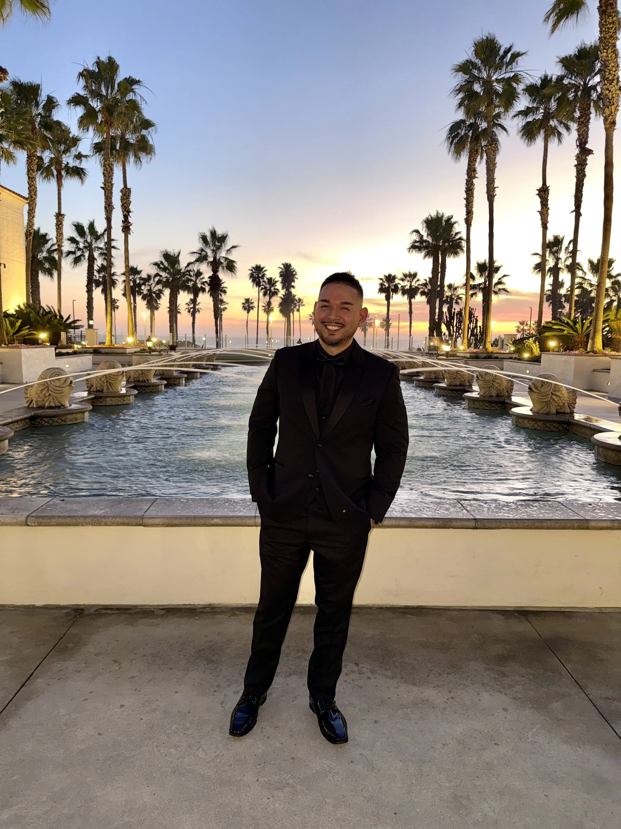 A man in a black tuxedo standing outdoors at sunset, smiling, with tall palm trees and a fountain in the background.