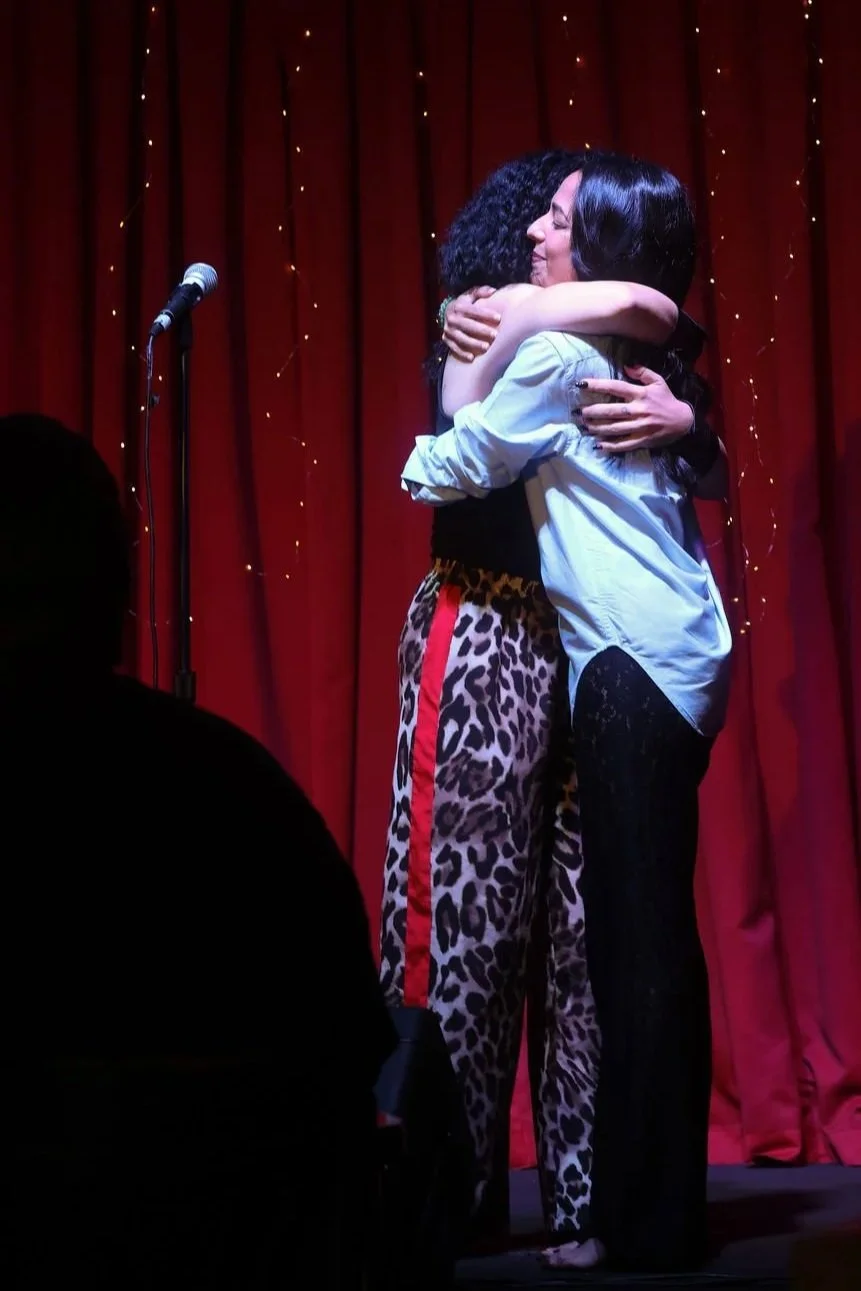 Two women hugging on stage in front of red curtain with small lights, one wearing leopard print pants and the other in a light-colored shirt.