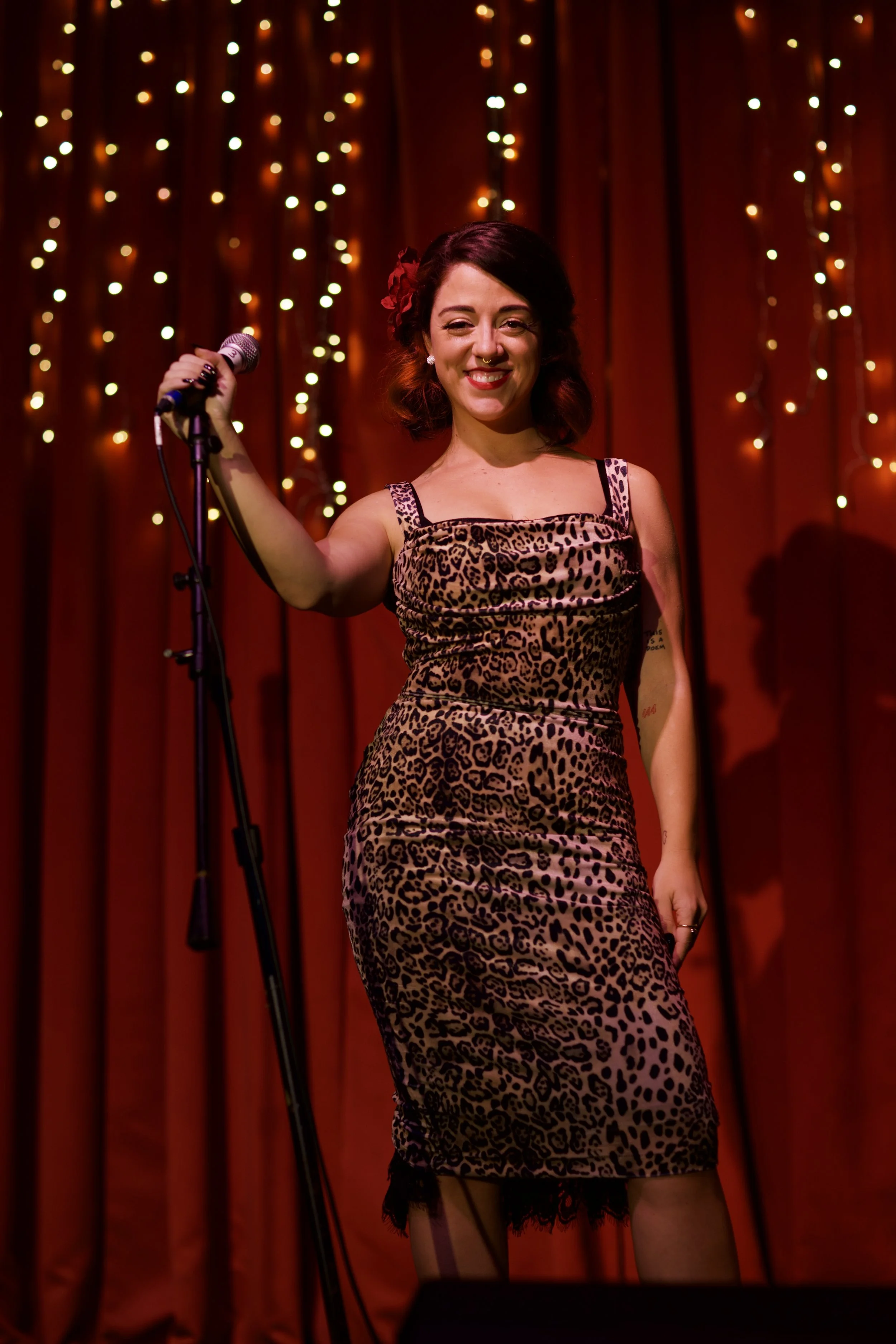 A woman in a leopard print dress standing on stage in front of red curtains with string lights, holding a microphone and smiling.
