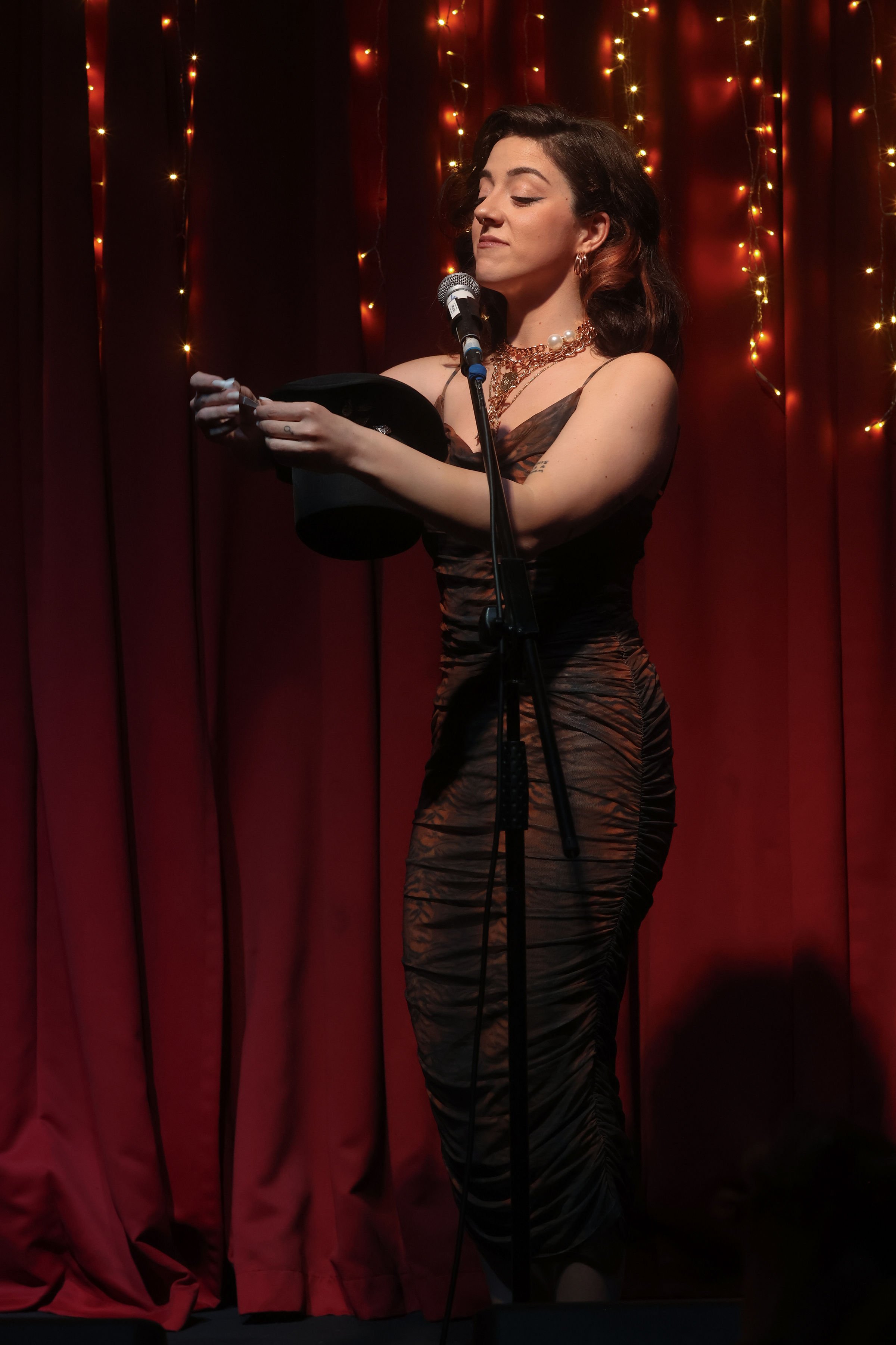 A woman standing on stage in front of a red curtain with string lights, holding a black hat and looking thoughtfully.