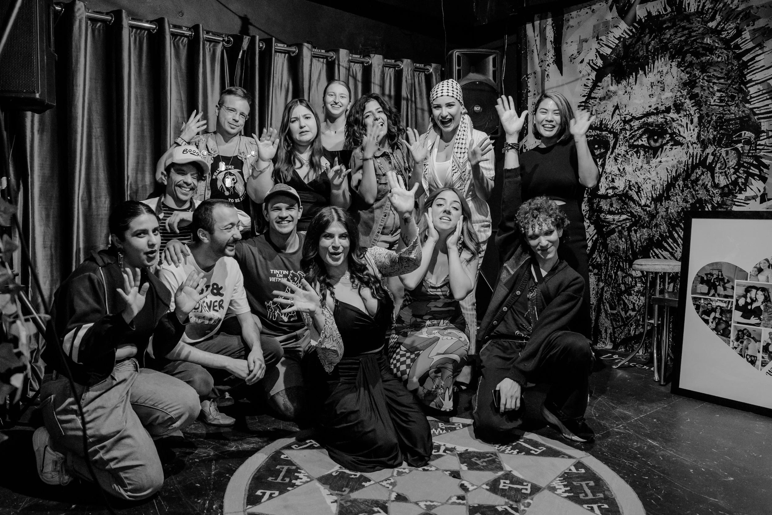 Group of nine people posing together in a dimly lit room with painted artwork on the wall, some sitting on the floor and some standing, all smiling and making playful gestures.