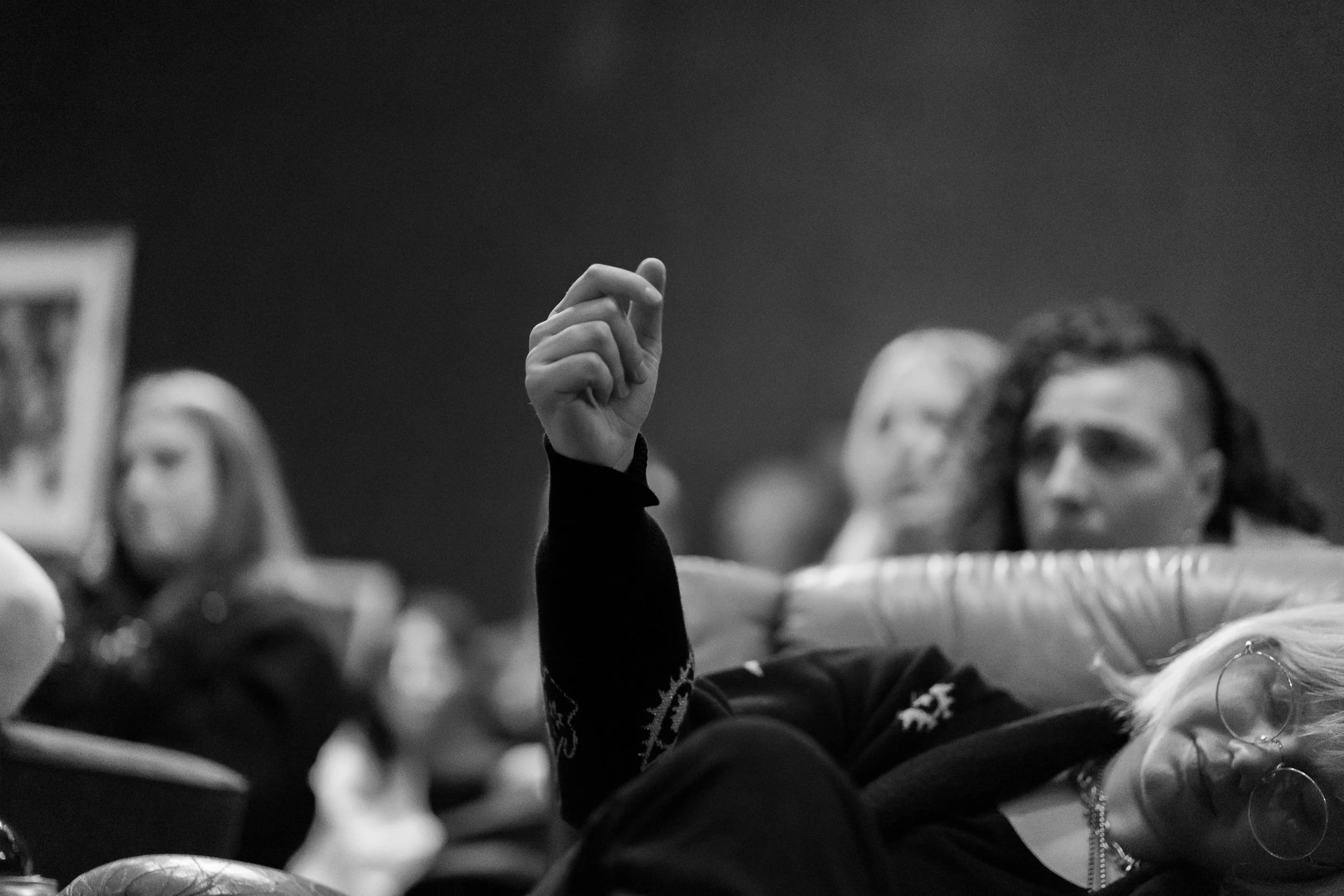 A black-and-white photo of a person raising their hand, clicking along to poetry in comraderie, among a crowd of seated people, with some blurry faces visible.