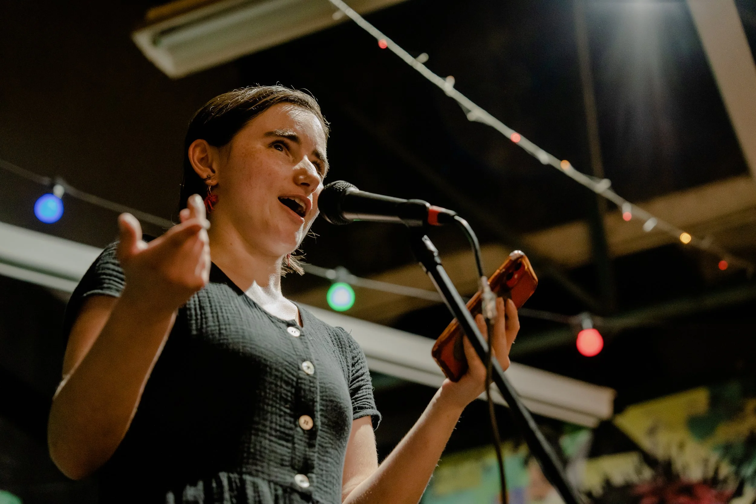 A woman singing into a microphone at an indoor event, holding a smartphone in her hand, with colorful string lights hanging in the background.