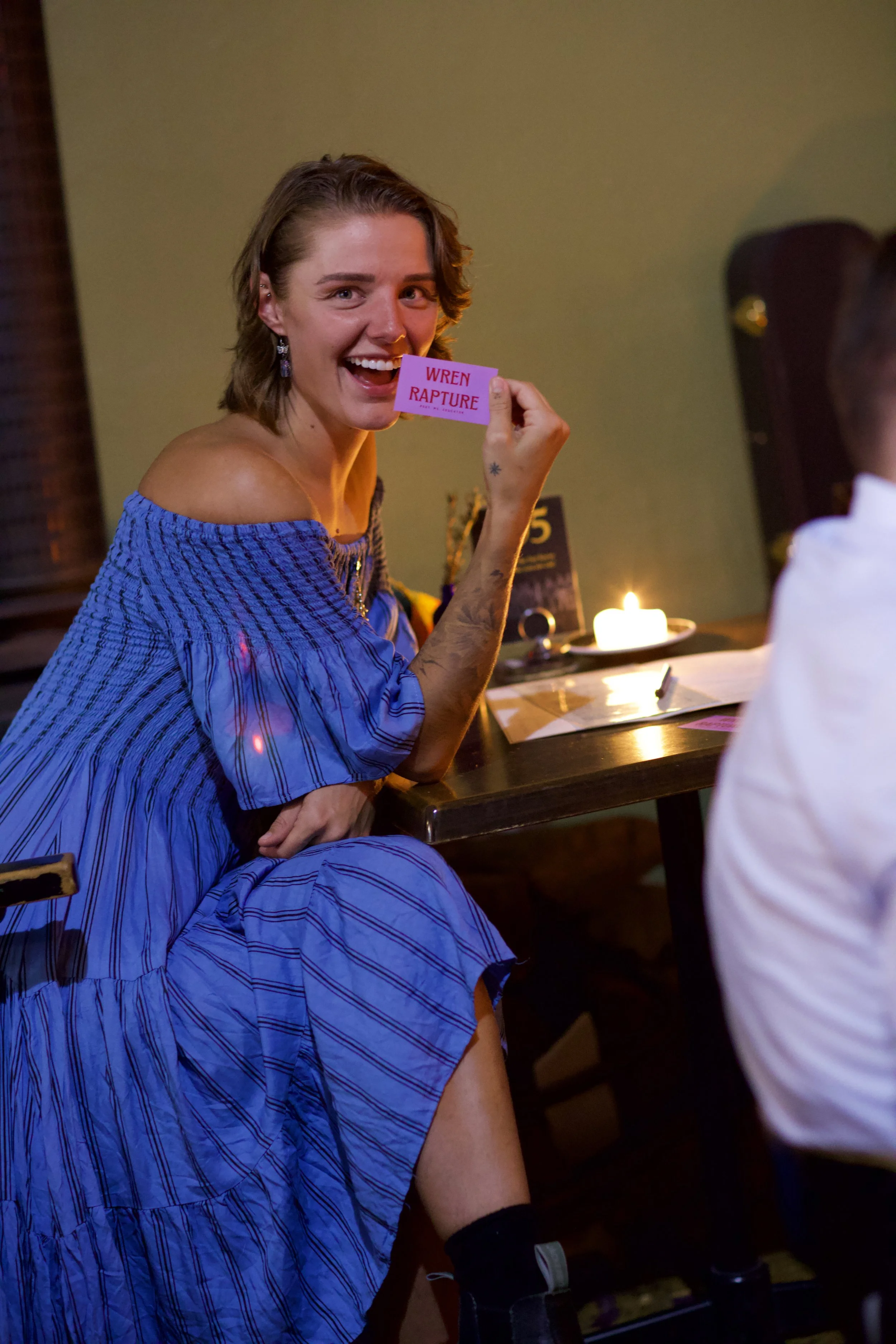 A woman with shoulder-length hair and earrings is smiling and holding a pink card that reads "WREN RAPTURE". She is wearing a blue and purple striped dress with off-the-shoulder sleeves, seated at a table with a lit candle and some papers in front of her.