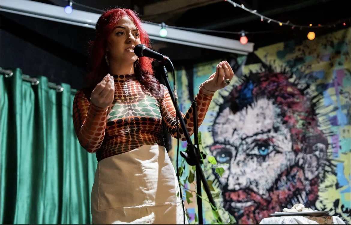 A woman with red hair and patterned top performs on stage with a microphone, in front of a colorful mural of a man's face with blue eyes.