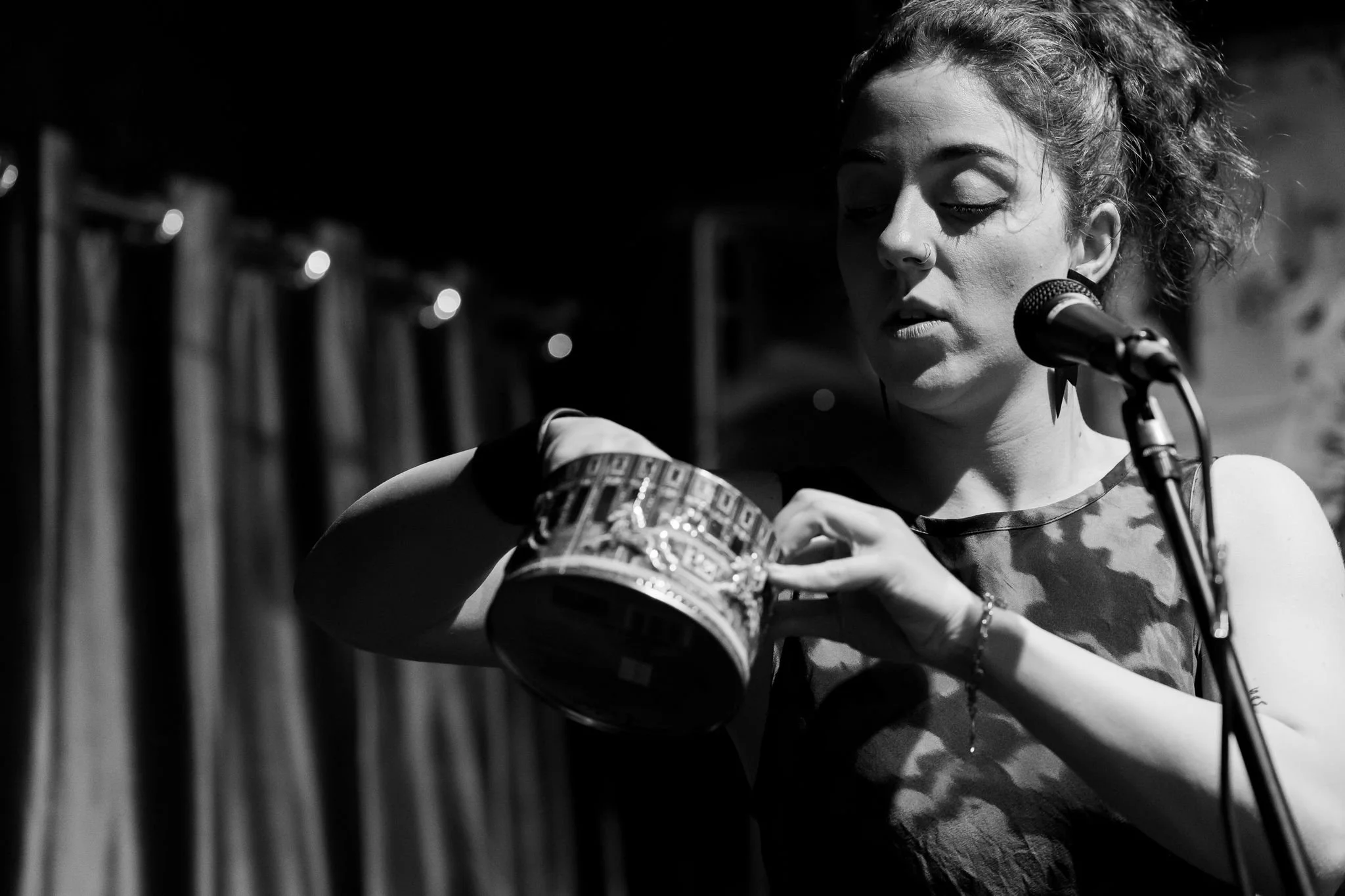 Black and white photo of a woman with curly hair and a nose piercing, looking down while holding a decorative cup, with a microphone in front of her.