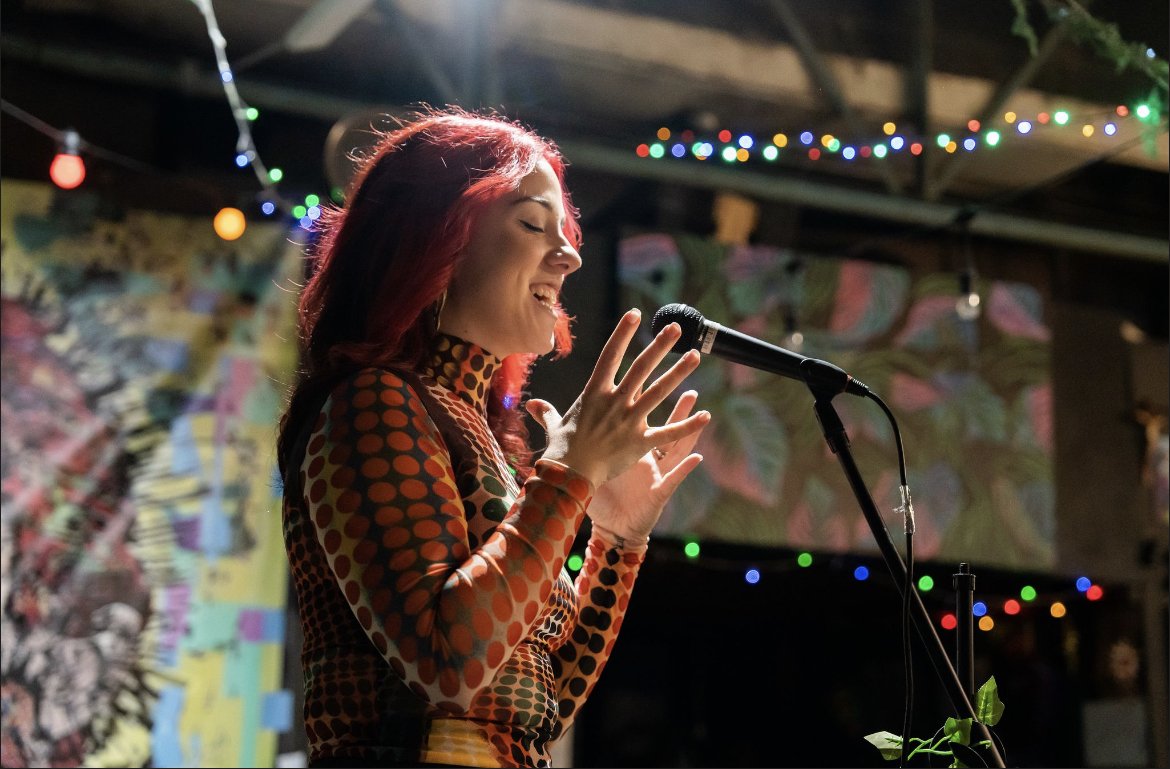 A woman with red hair singing into a microphone at a decorated venue with colorful string lights and a vibrant mural background.