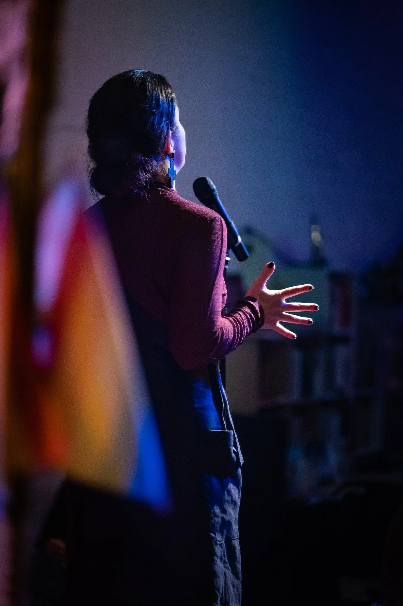 A woman with dark hair, wearing a red top and large blue earrings, stands on stage in front of a microphone, gesturing with her right hand, while speaking or singing in a dark, dimly-lit environment with stage lighting.