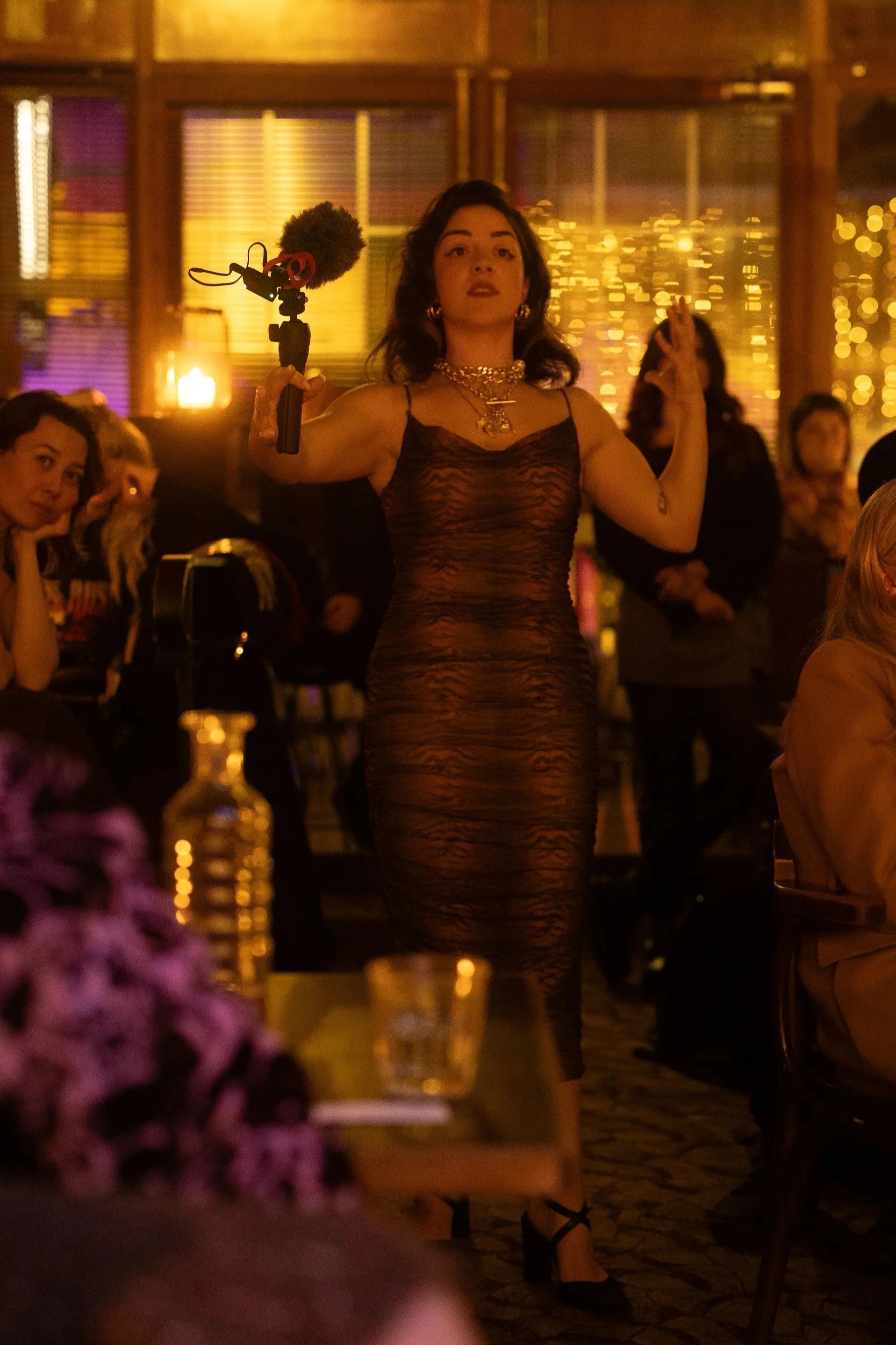 A woman in a tight, tiger-stripe dress holding a microphone on a pole at a lively indoor event with people seated at tables, warm lighting, and decorative string lights in the background.