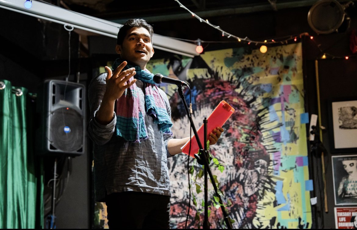 A man with dark hair, wearing a gray shirt and colorful scarf, speaking into a microphone on a stage decorated with colorful, abstract background art and string lights.