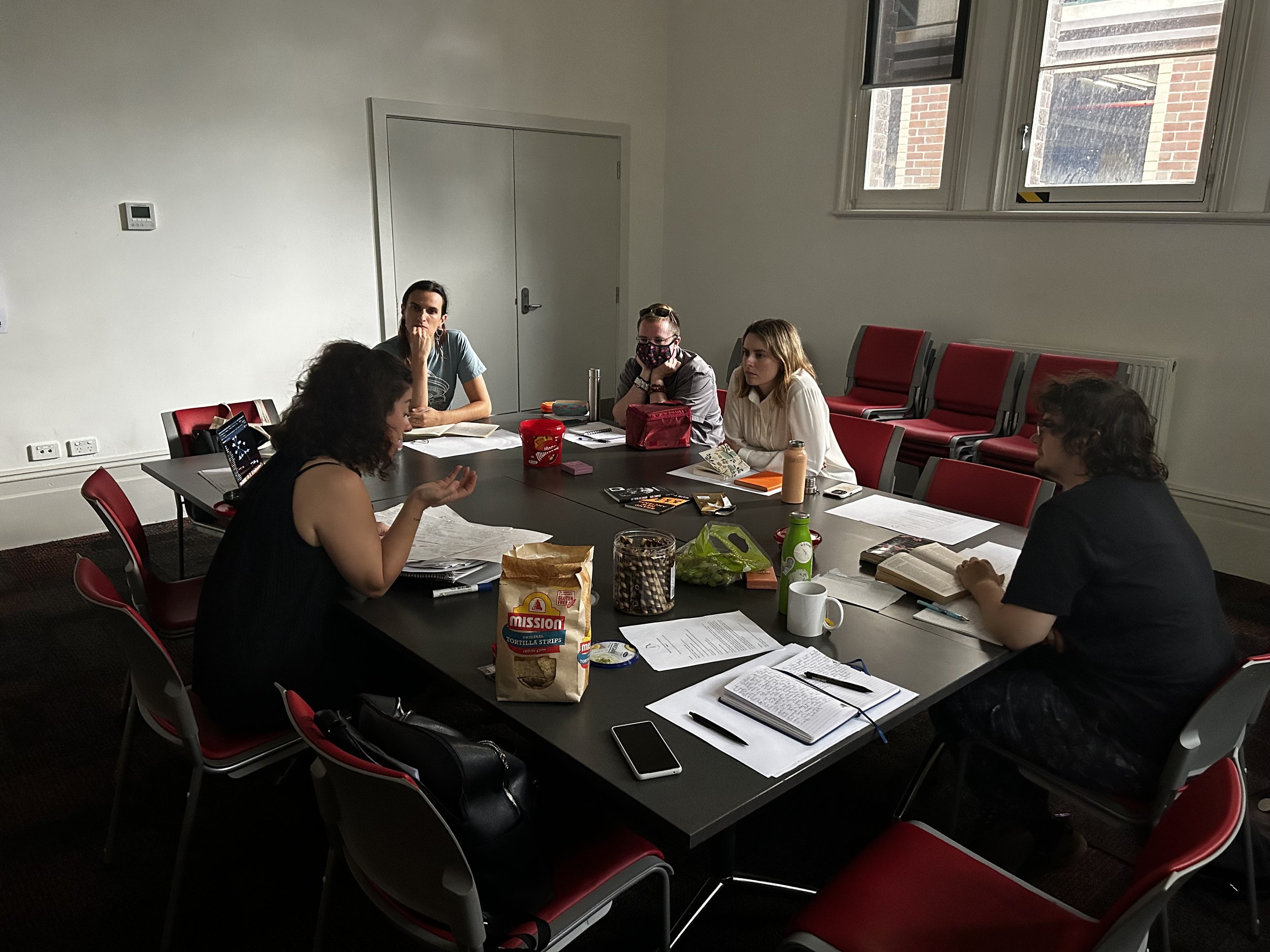 A group of five people sitting around a table in a meeting room. There are notes, drinks, and snacks on the table. Large windows let in natural light, and red chairs are positioned around the table and along the wall.