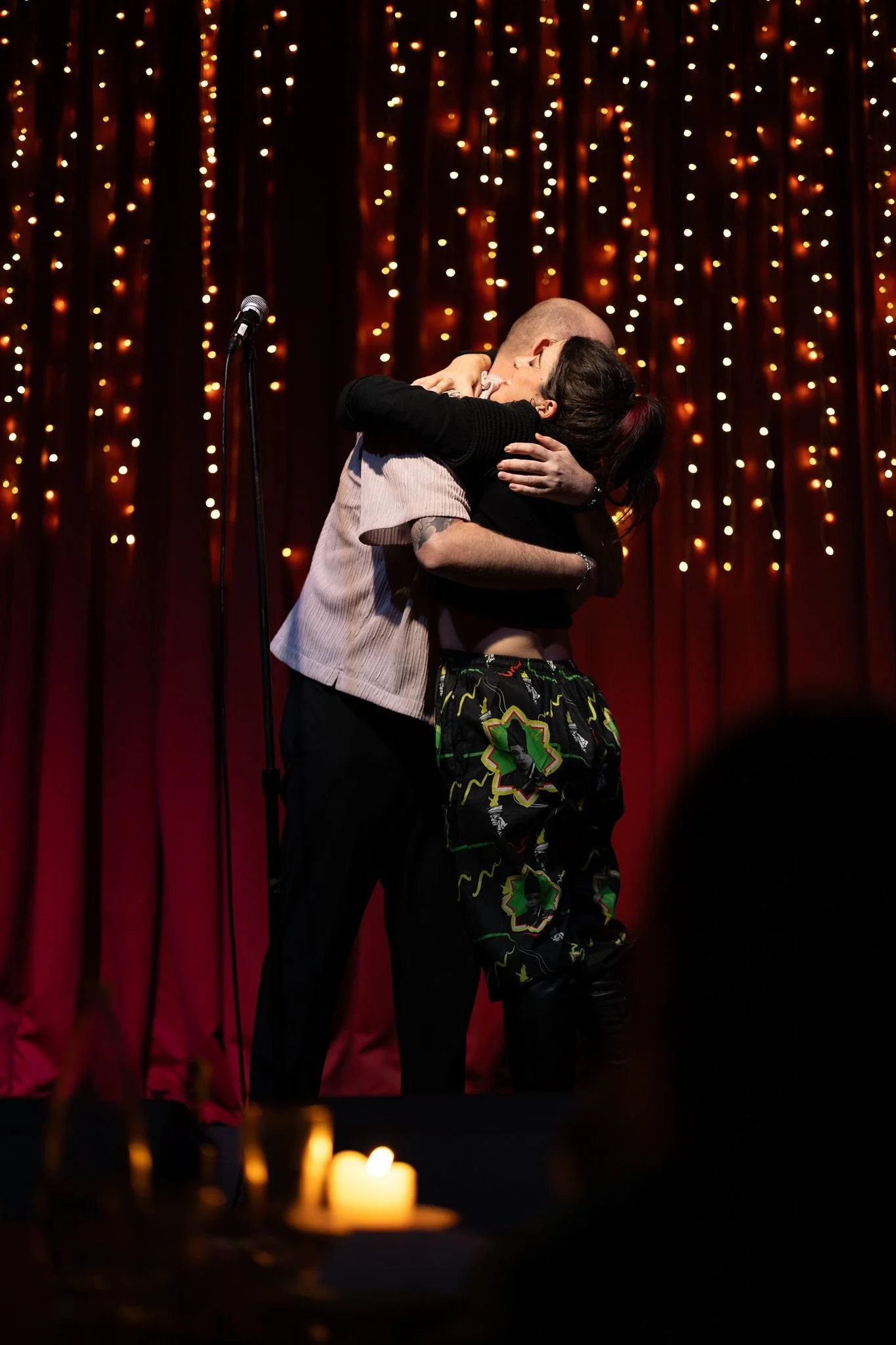 A couple of friends hugging in celebration on stage in front of a backdrop of string lights, with candles in the foreground.