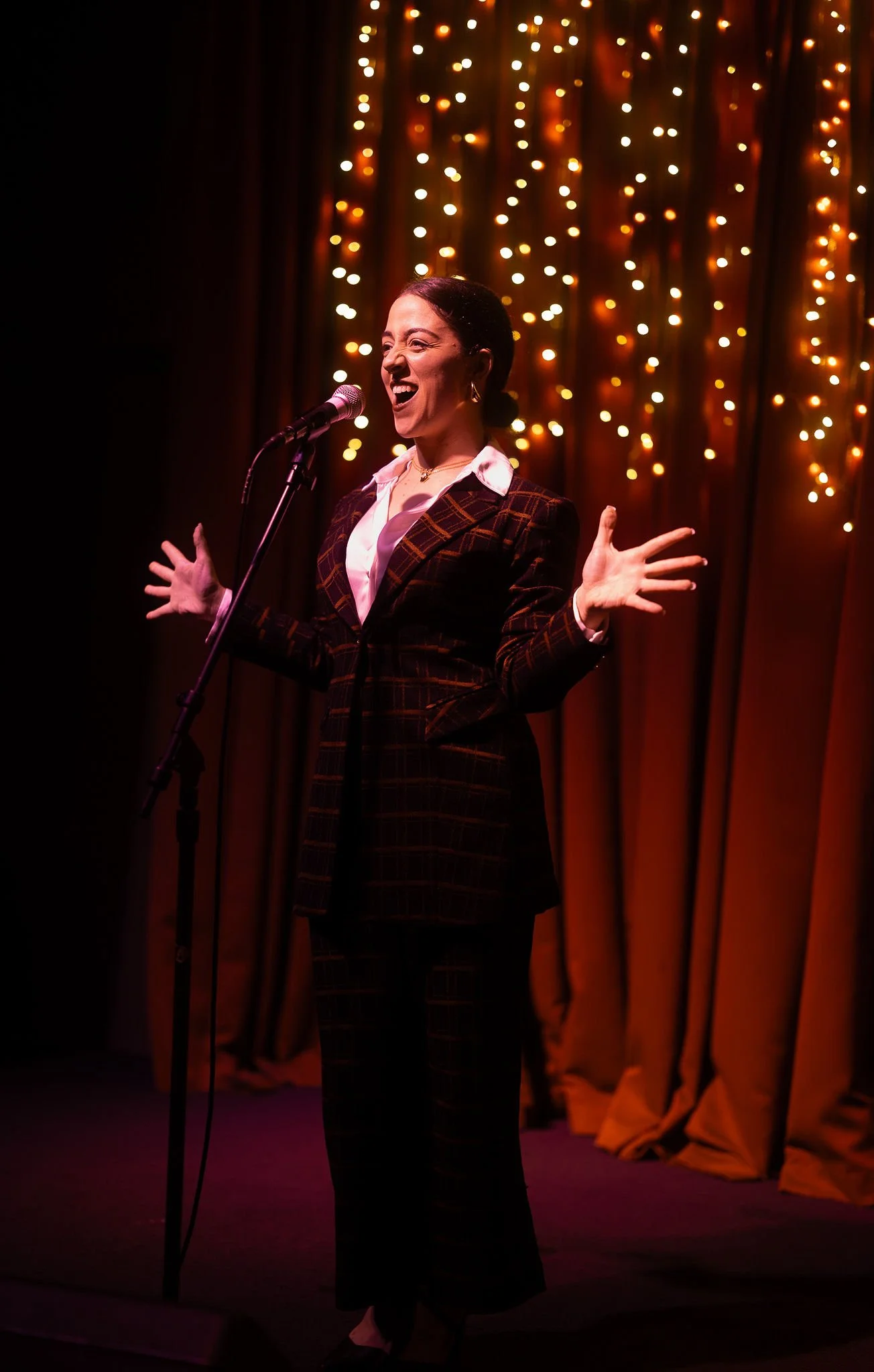 Woman singing or speaking into a microphone on stage with string lights and orange curtains in the background. She is smiling and gesturing with open hands.