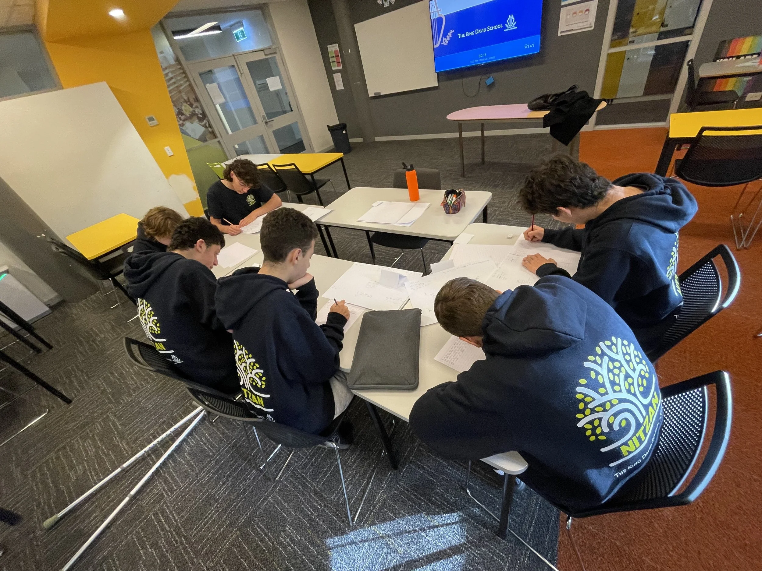 Students sitting at tables in a classroom, writing and studying with papers and notebooks, and a large screen on the wall displaying 'The King David School' logo.