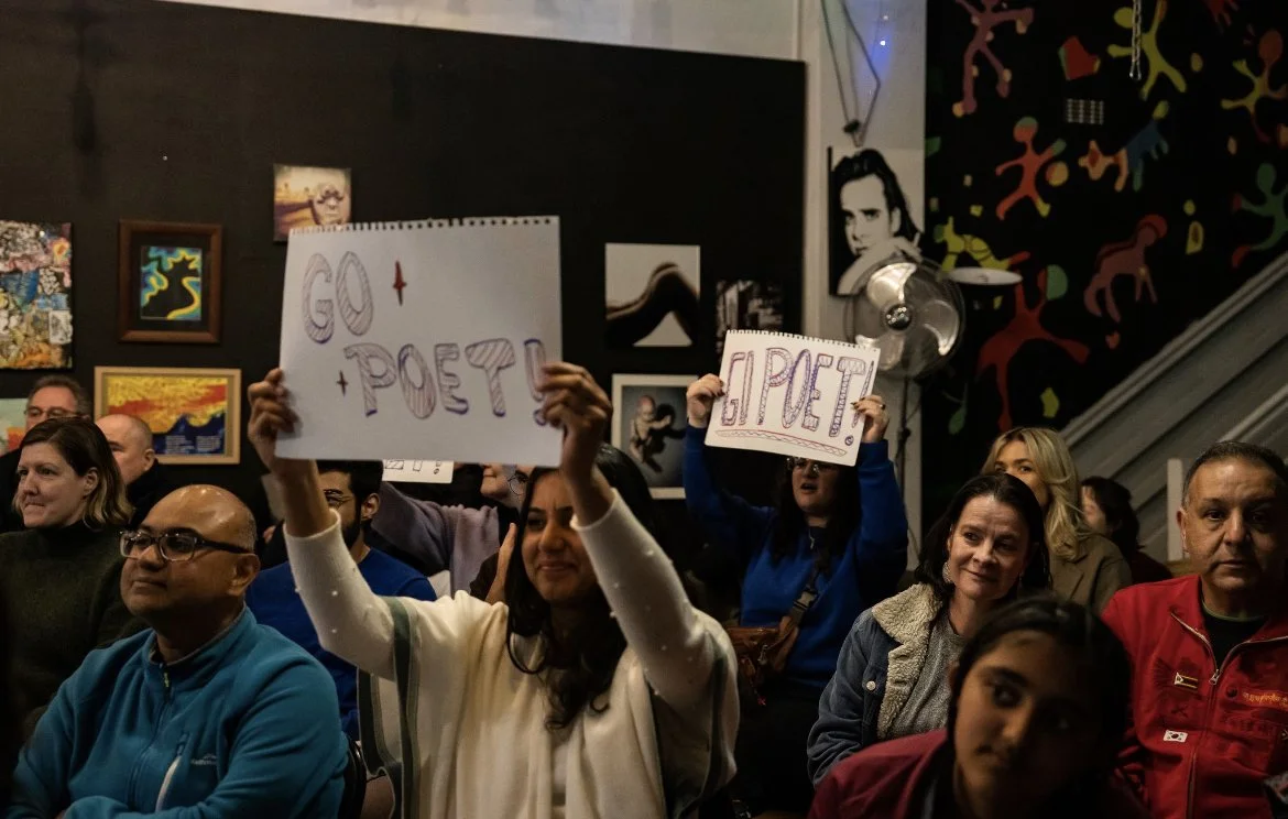 People attending a cultural event, some holding signs that say 'Go Poet' and 'Hi poet,' in a room with colorful artwork on the walls.