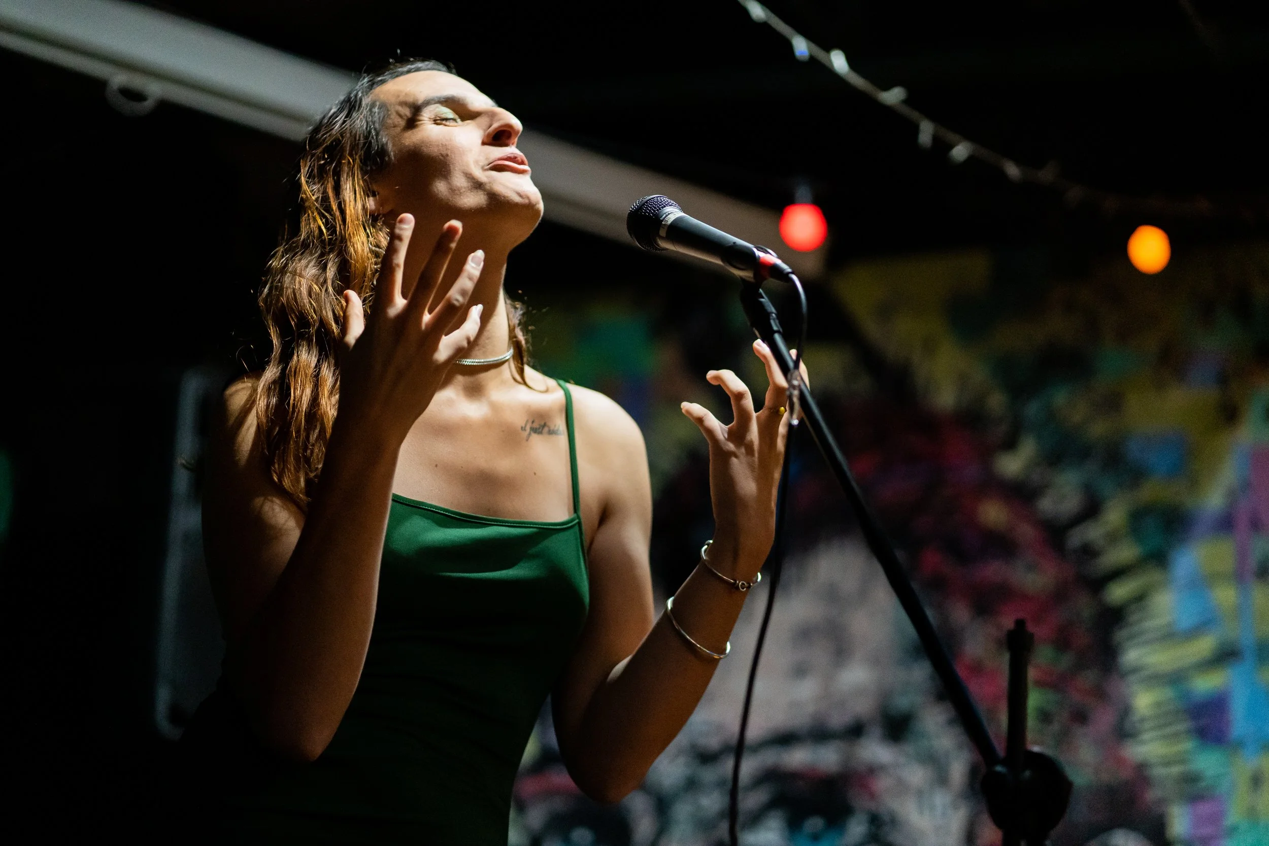 A woman with long, wavy hair wearing a green spaghetti strap top is singing passionately into a microphone on stage with her eyes closed and hands raised.