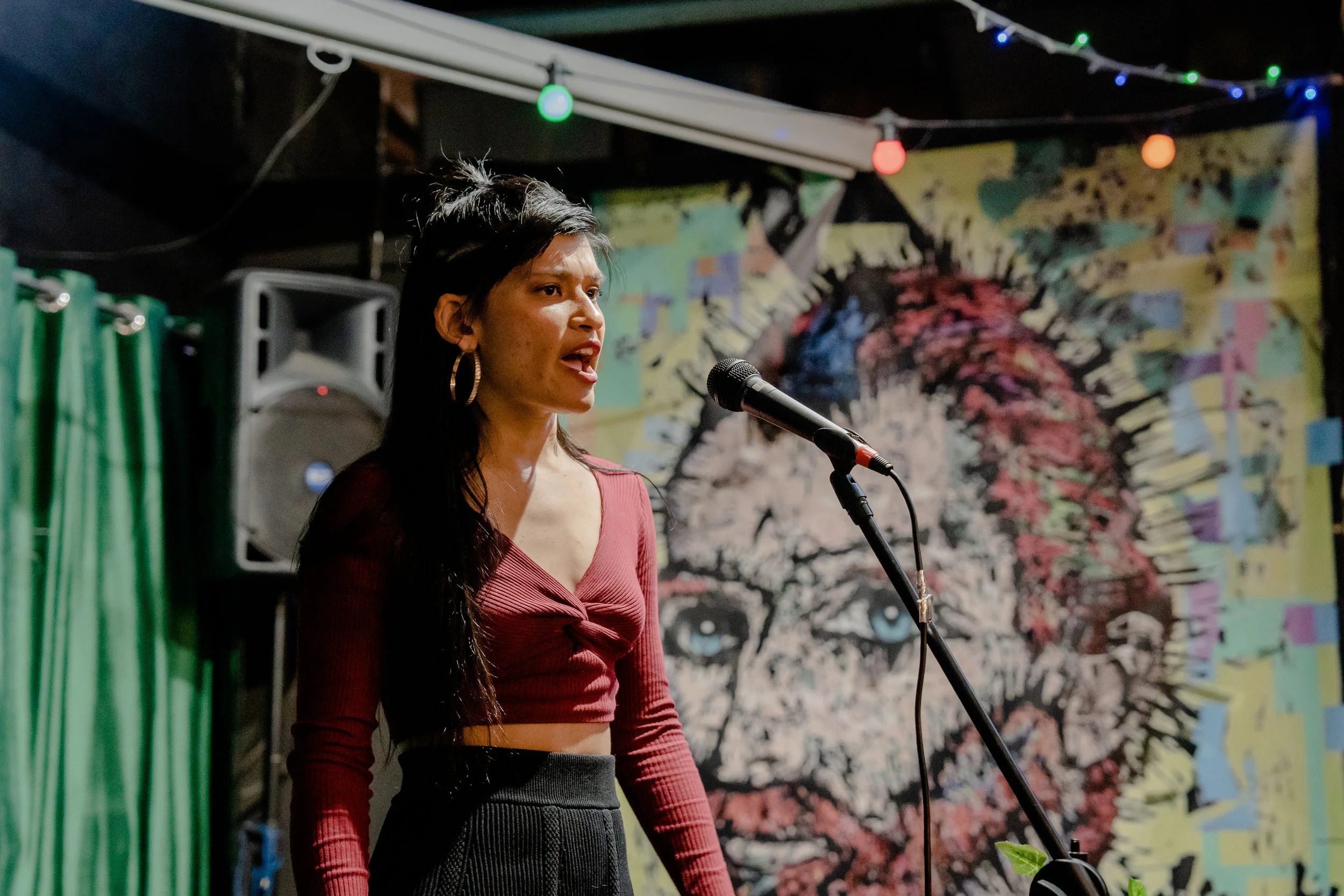 A woman with long dark hair and hoop earrings speaking into a microphone at an indoor event with colorful murals and string lights in the background.