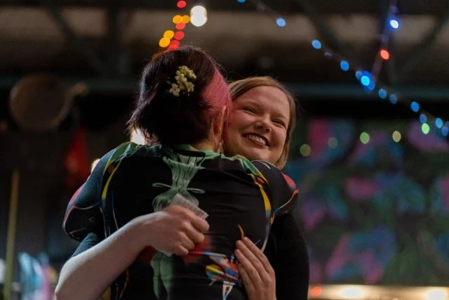Two women hugging and smiling at a party or celebration under colorful string lights.