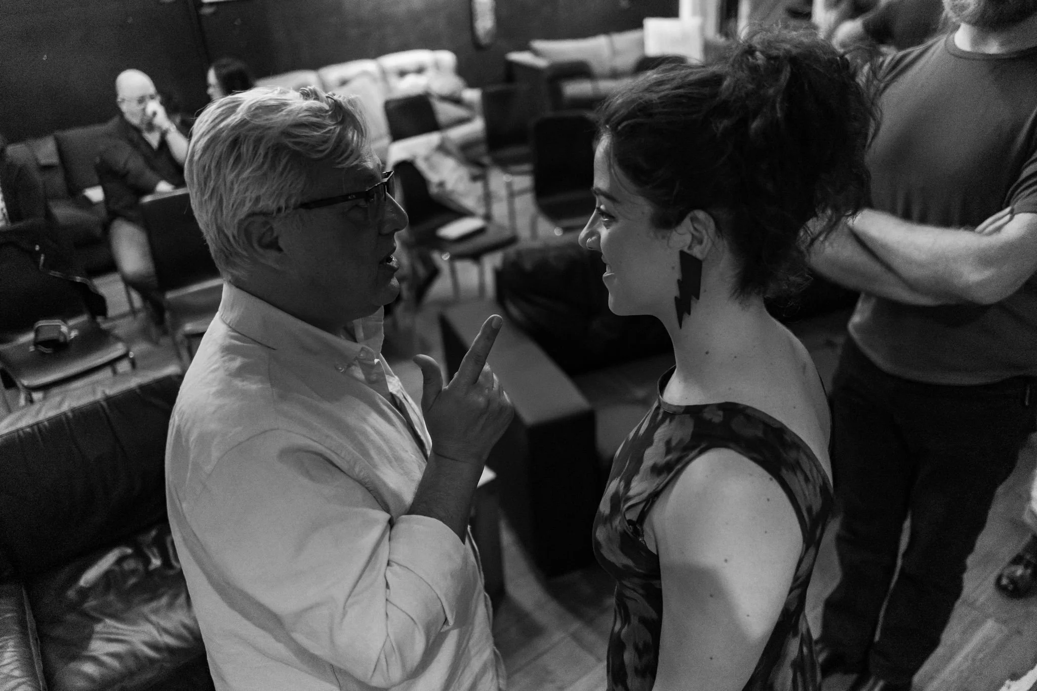 A man and a woman face each other in close proximity, engaging in an intense conversation or debate. The man has light-colored hair and glasses, wearing a white shirt, and is gesturing with his finger. The woman has dark, curly hair, earrings, and a 